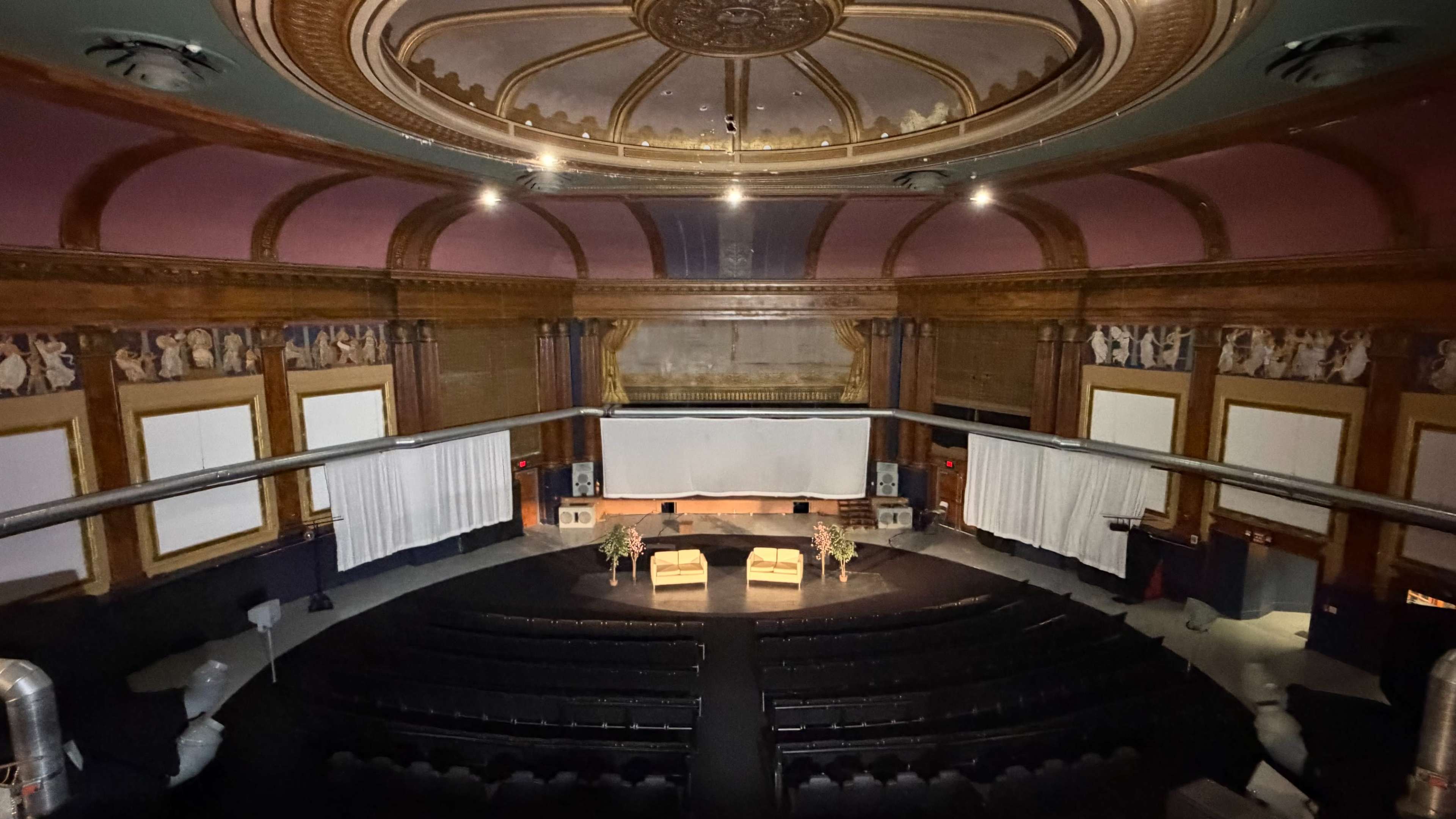 The image shows an ornate theater interior with a circular ceiling, rows of seats, and a large screen set up on stage.