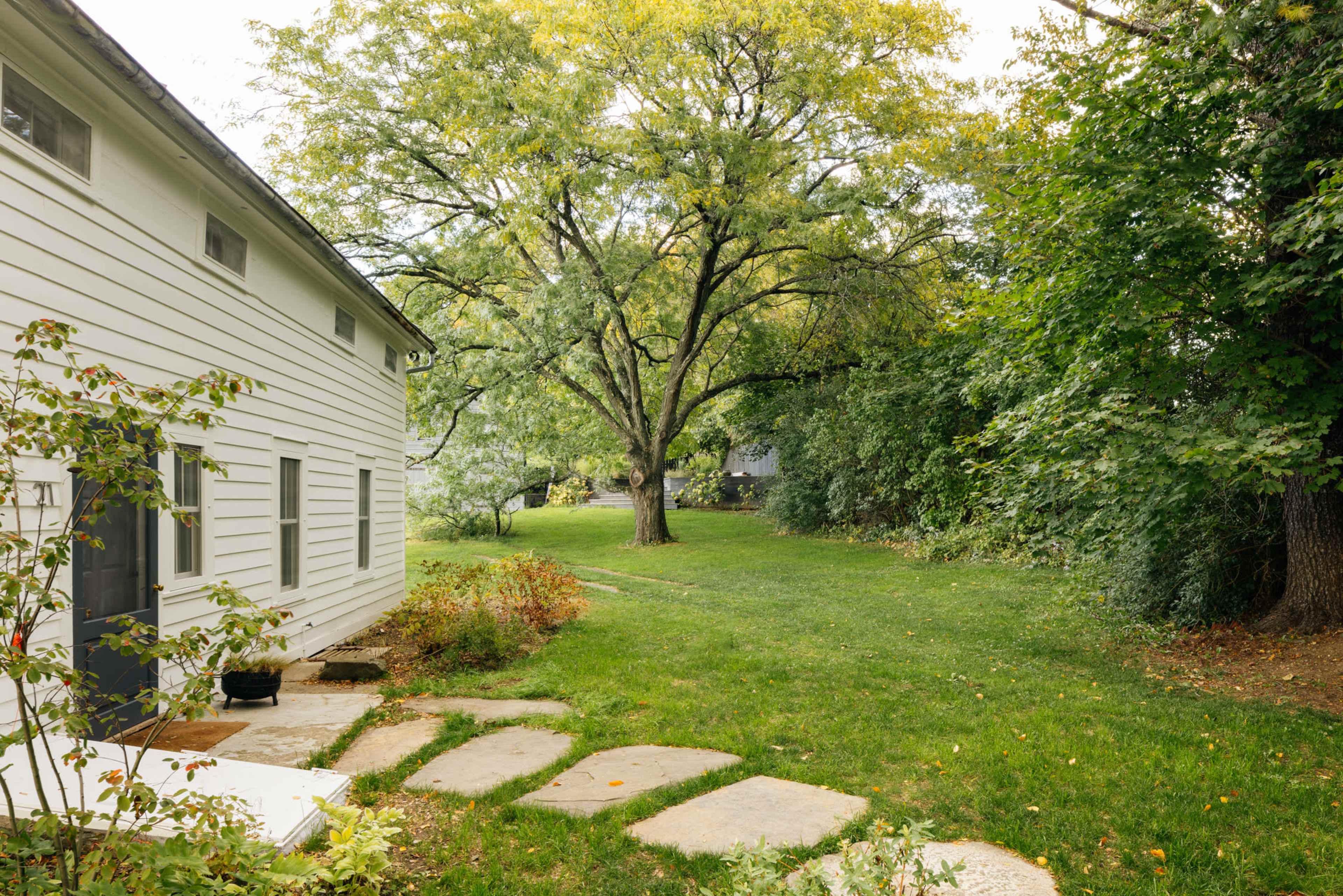 A well-maintained white house sits beside a grassy yard with a large tree and a stone pathway leading to it.