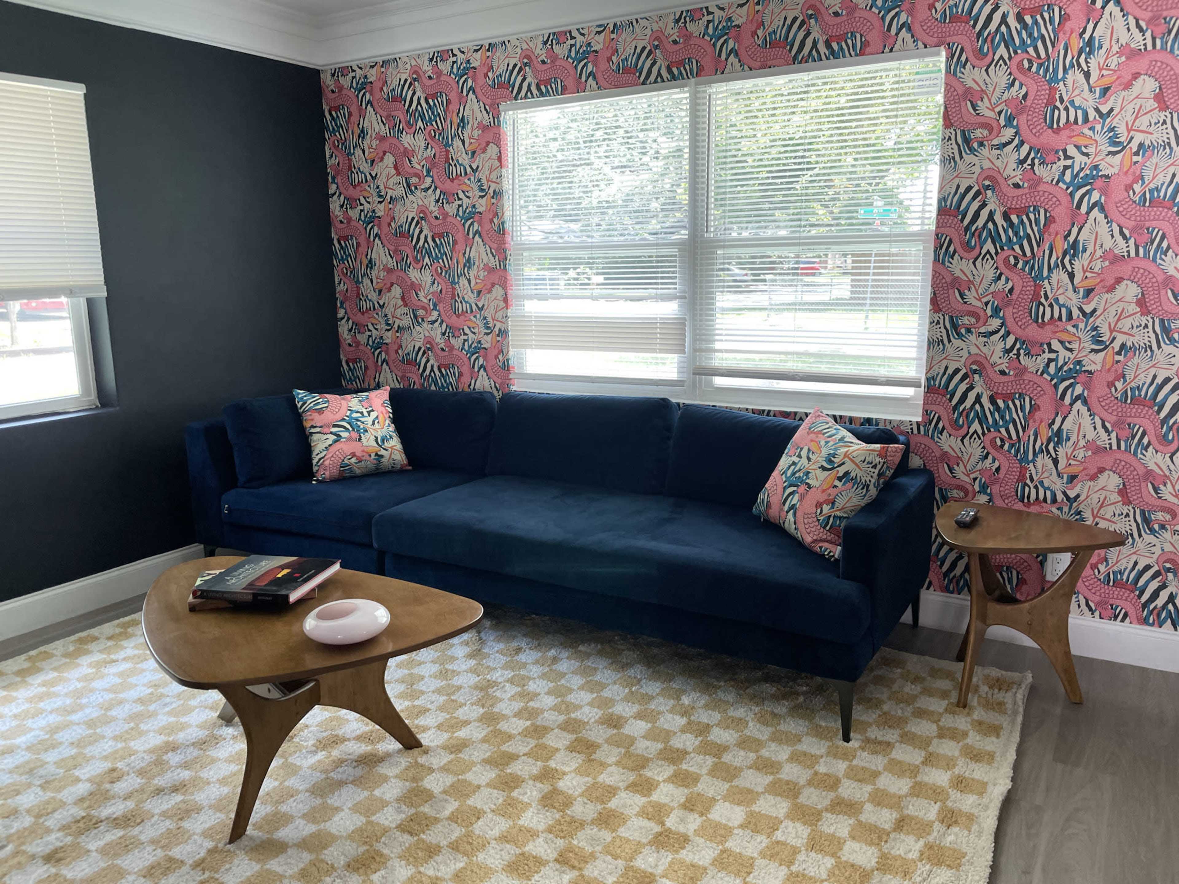 The image shows a modern living room featuring a dark blue sectional sofa with patterned throw pillows, a wooden side table, and a patterned wall with palm leaf designs.