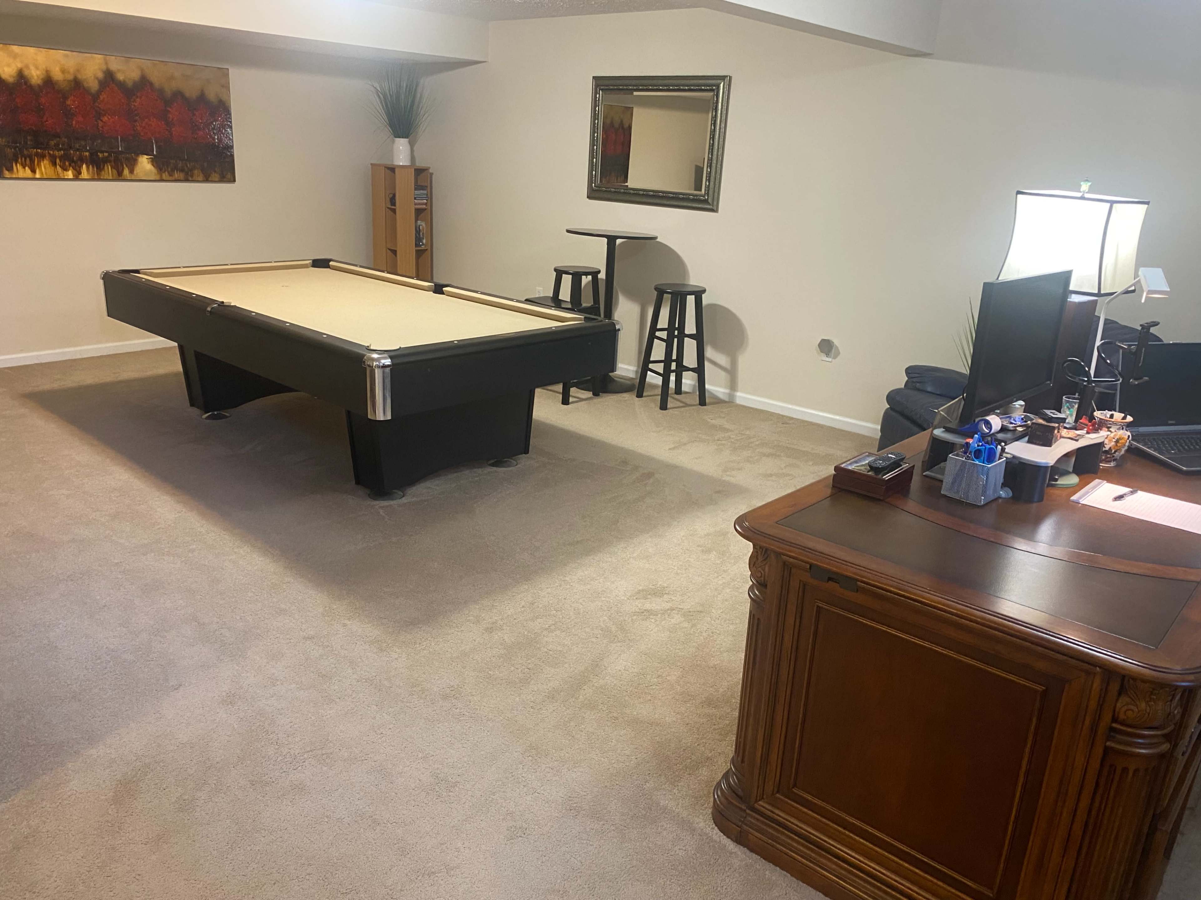 The image shows a spacious basement room featuring a pool table in the center, a desk with a computer setup to the right, and a few seating options along with a decorative mirror on the wall.