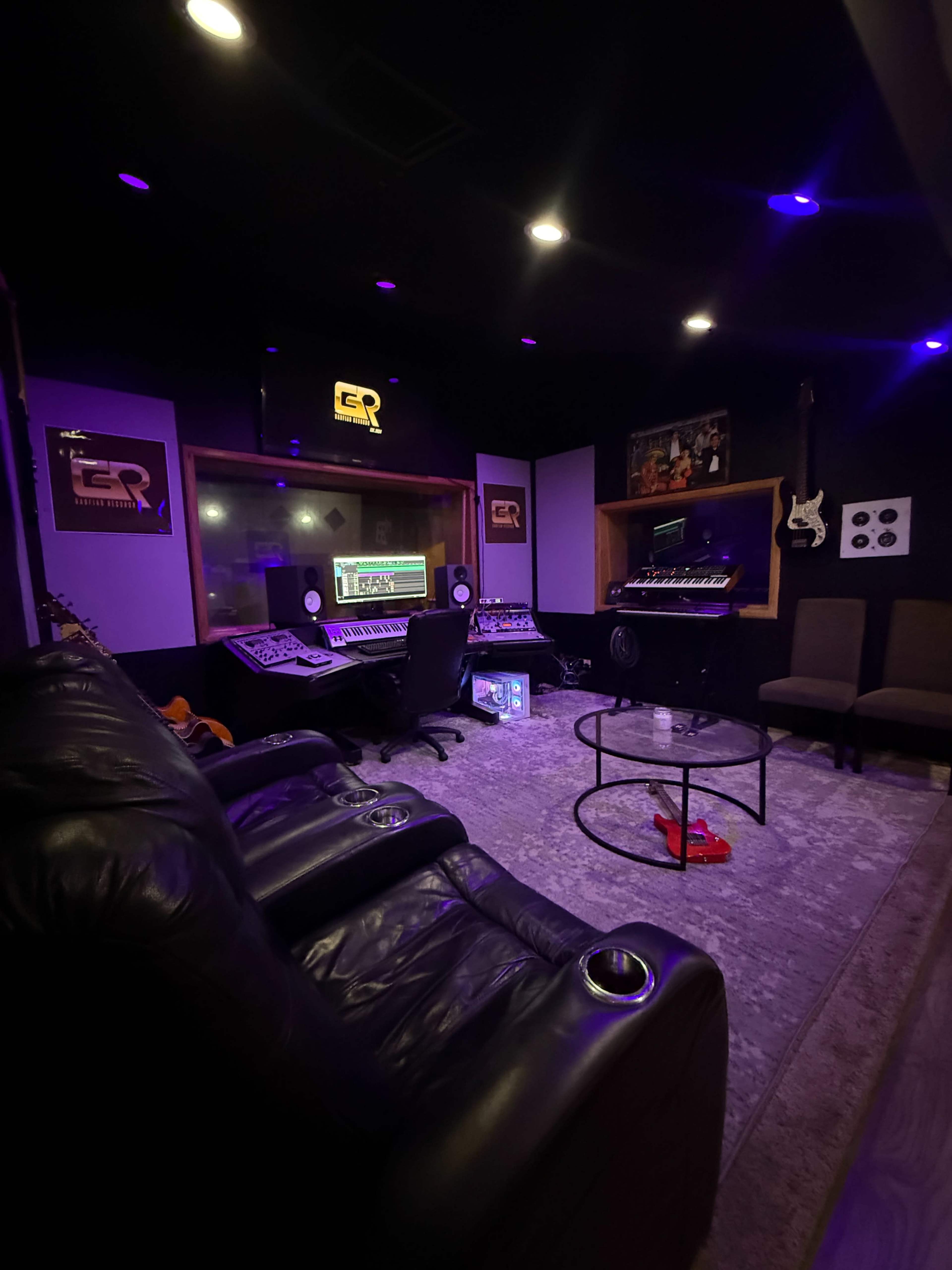 The image shows a modern recording studio with a dark color scheme, featuring a control desk, synthesizers, and comfortable seating areas.