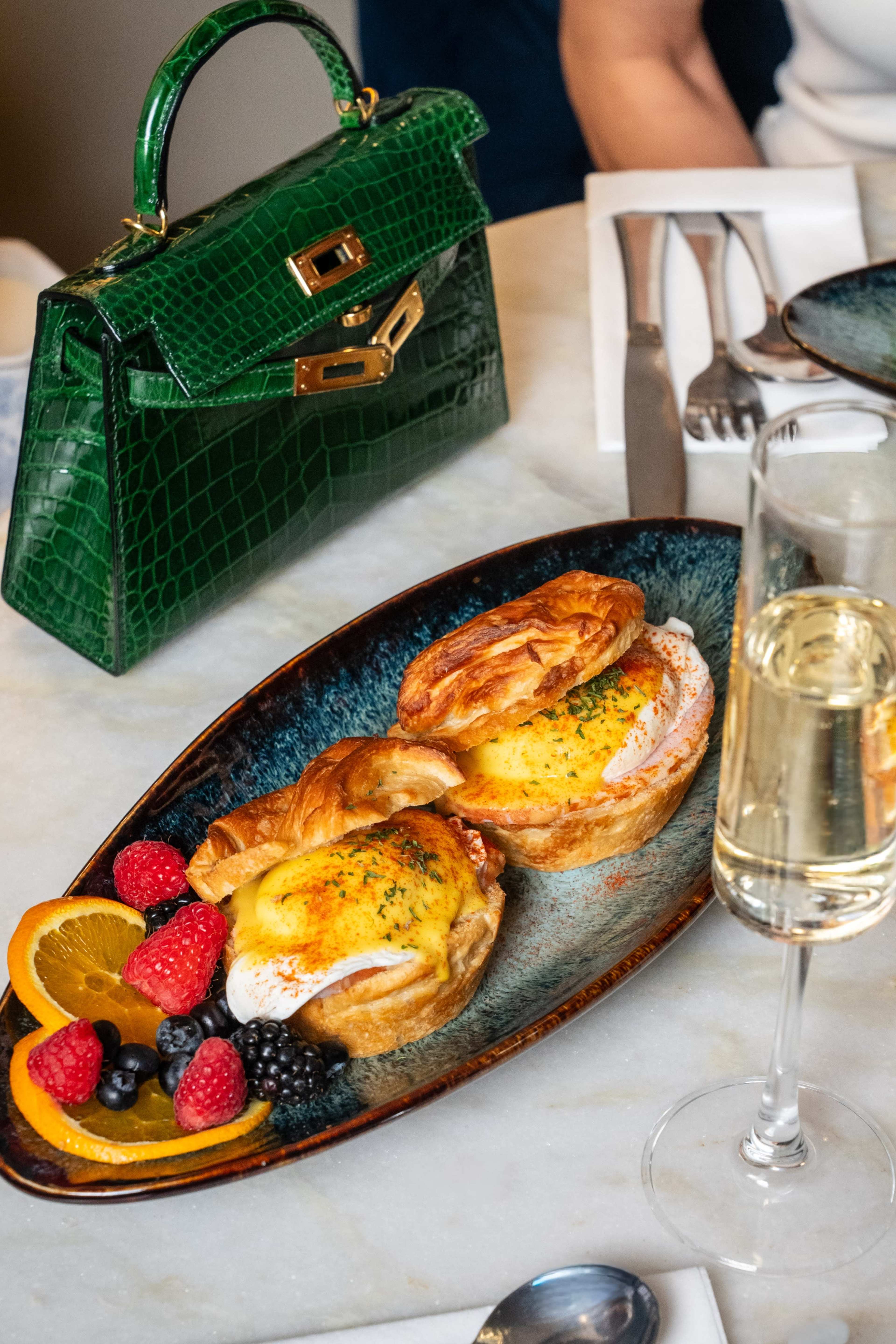 A plate holds two baked egg dishes alongside an assortment of fresh fruit, while a stylish green handbag is positioned nearby on the table.