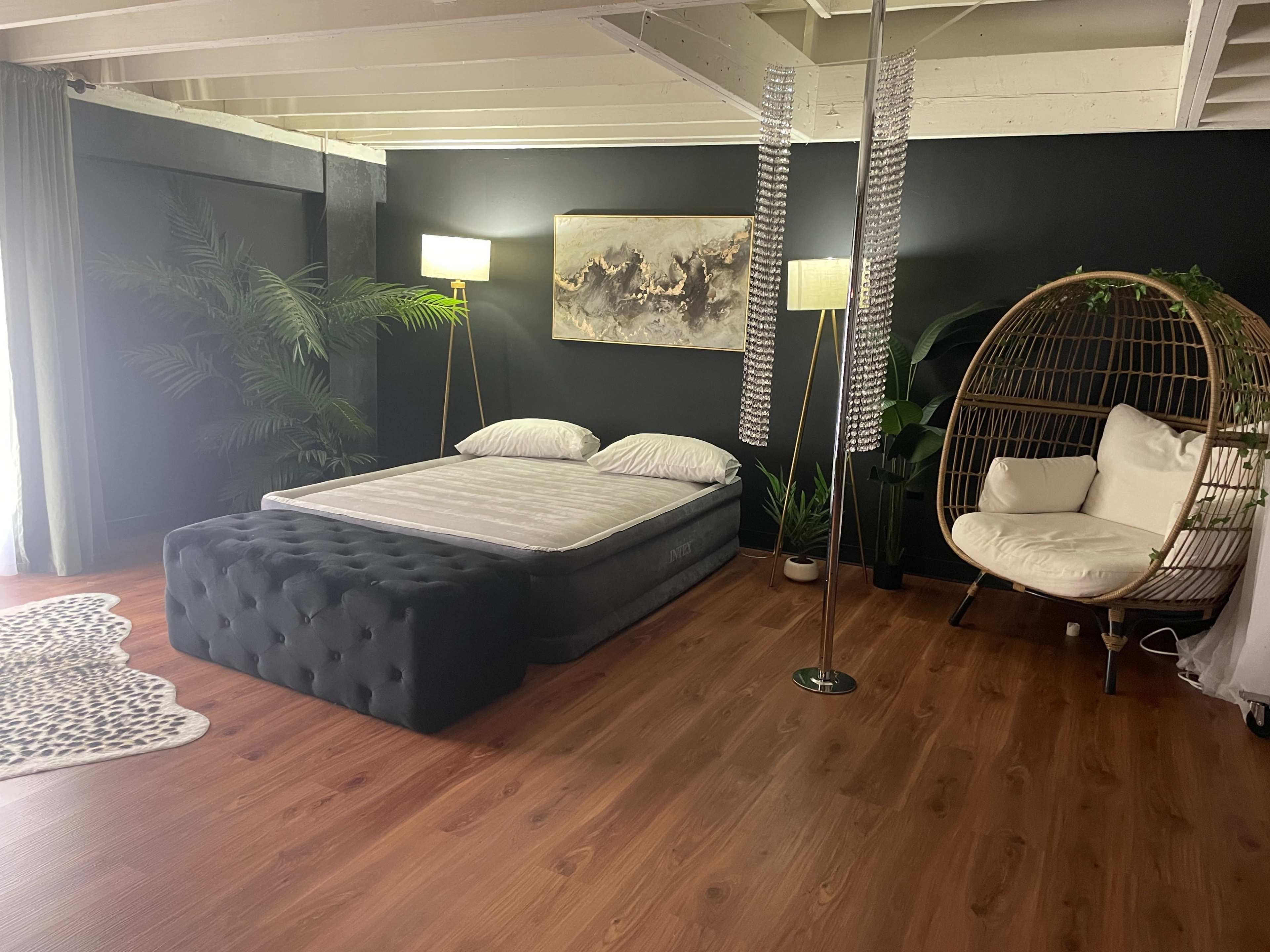 The image shows a modern bedroom with a bed, a cozy seating area in a chair, and decorative plants against a dark wall.