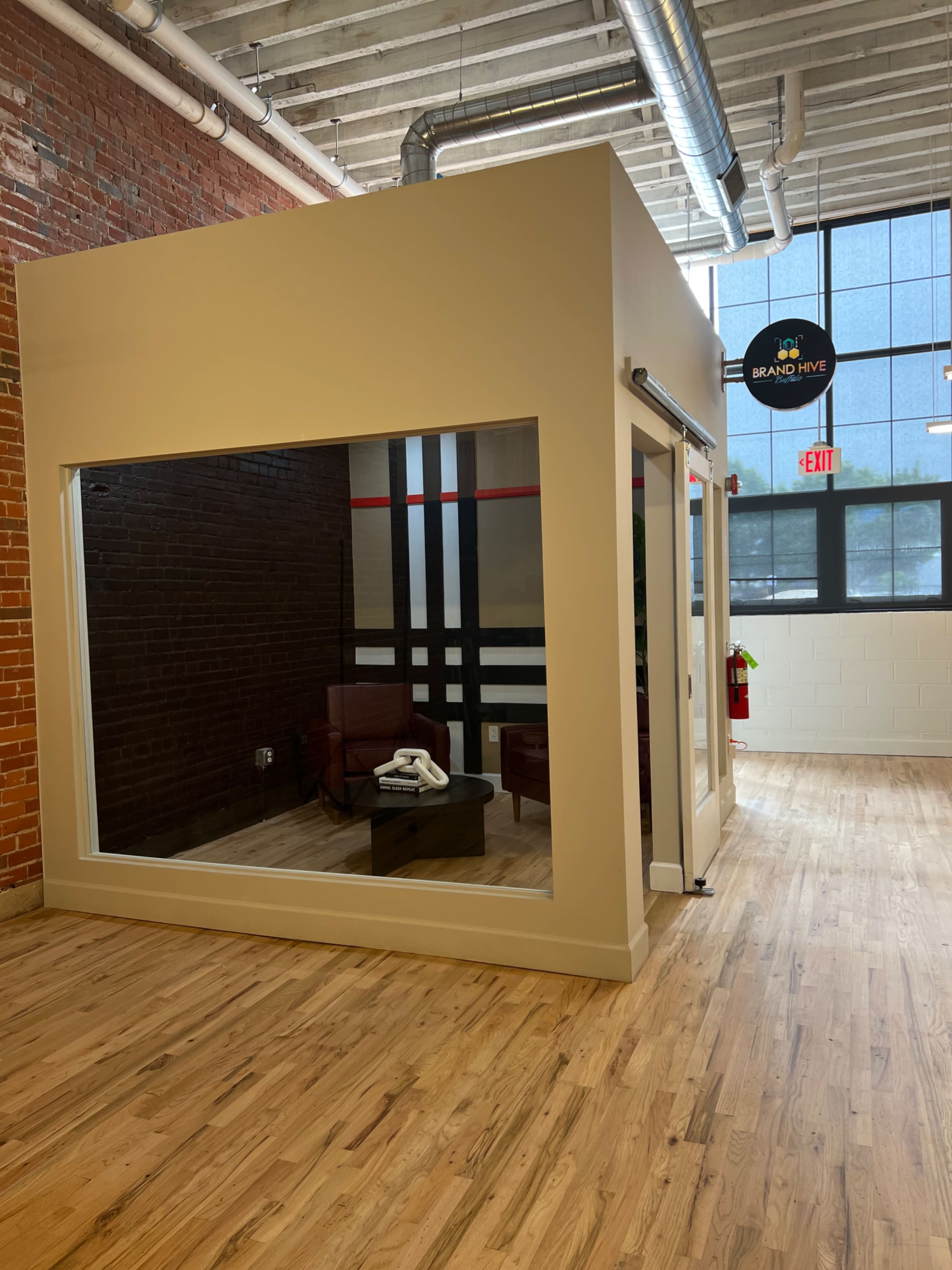 A modern glass-walled office cubicle with a wooden floor and an exposed brick wall.