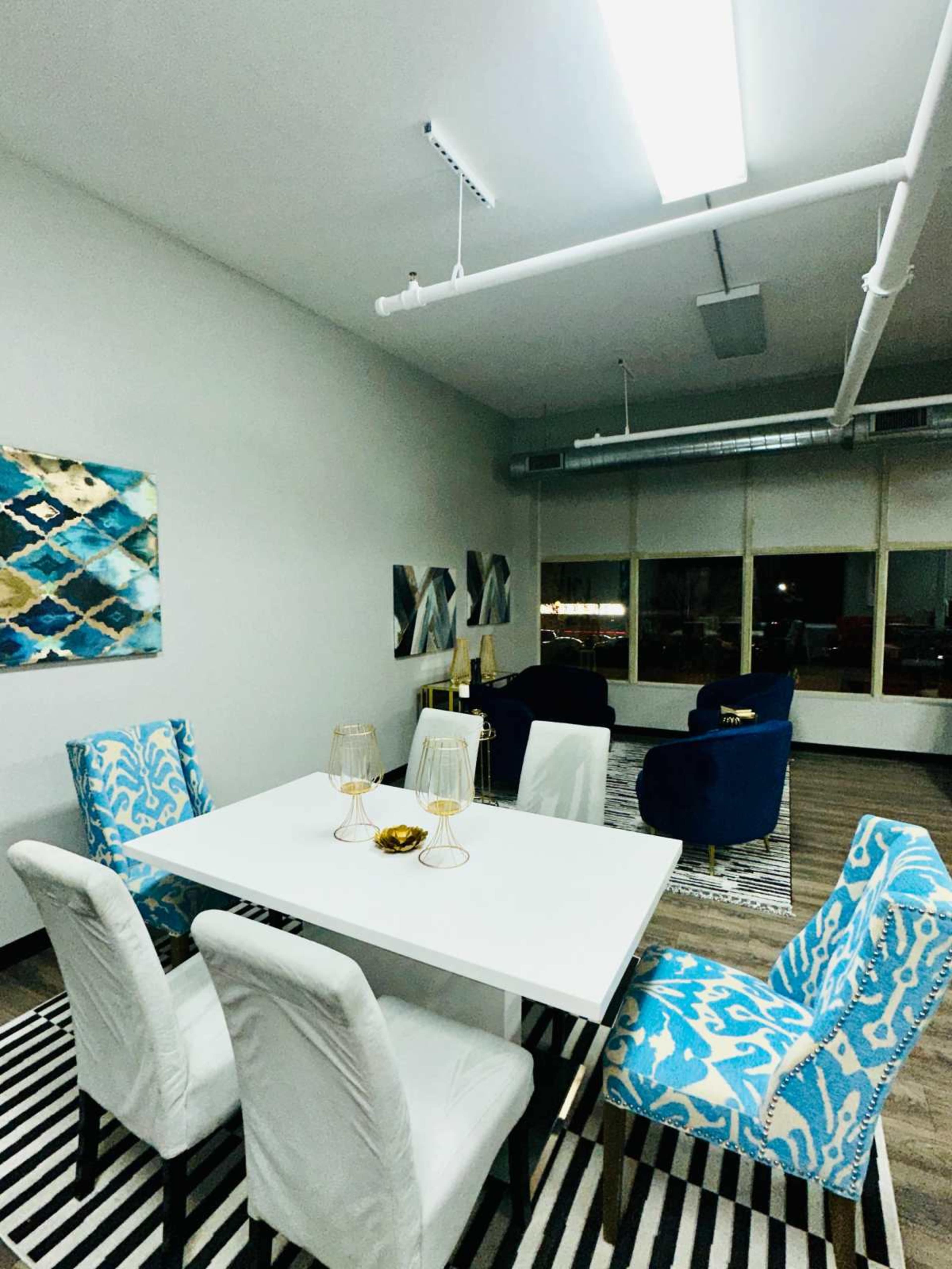 A modern dining area features a white table surrounded by blue and white patterned chairs, with abstract artwork on the walls and a striped rug on the floor.