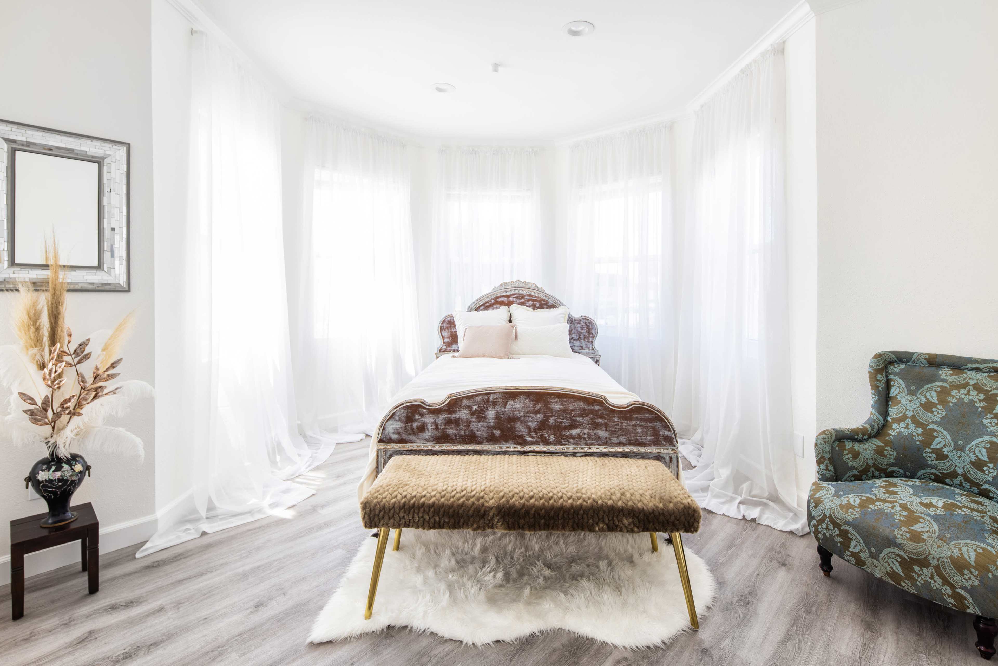 A bright bedroom featuring a bed with a patterned headboard, a decorative bench at the foot, and sheer curtains allowing sunlight to filter in.