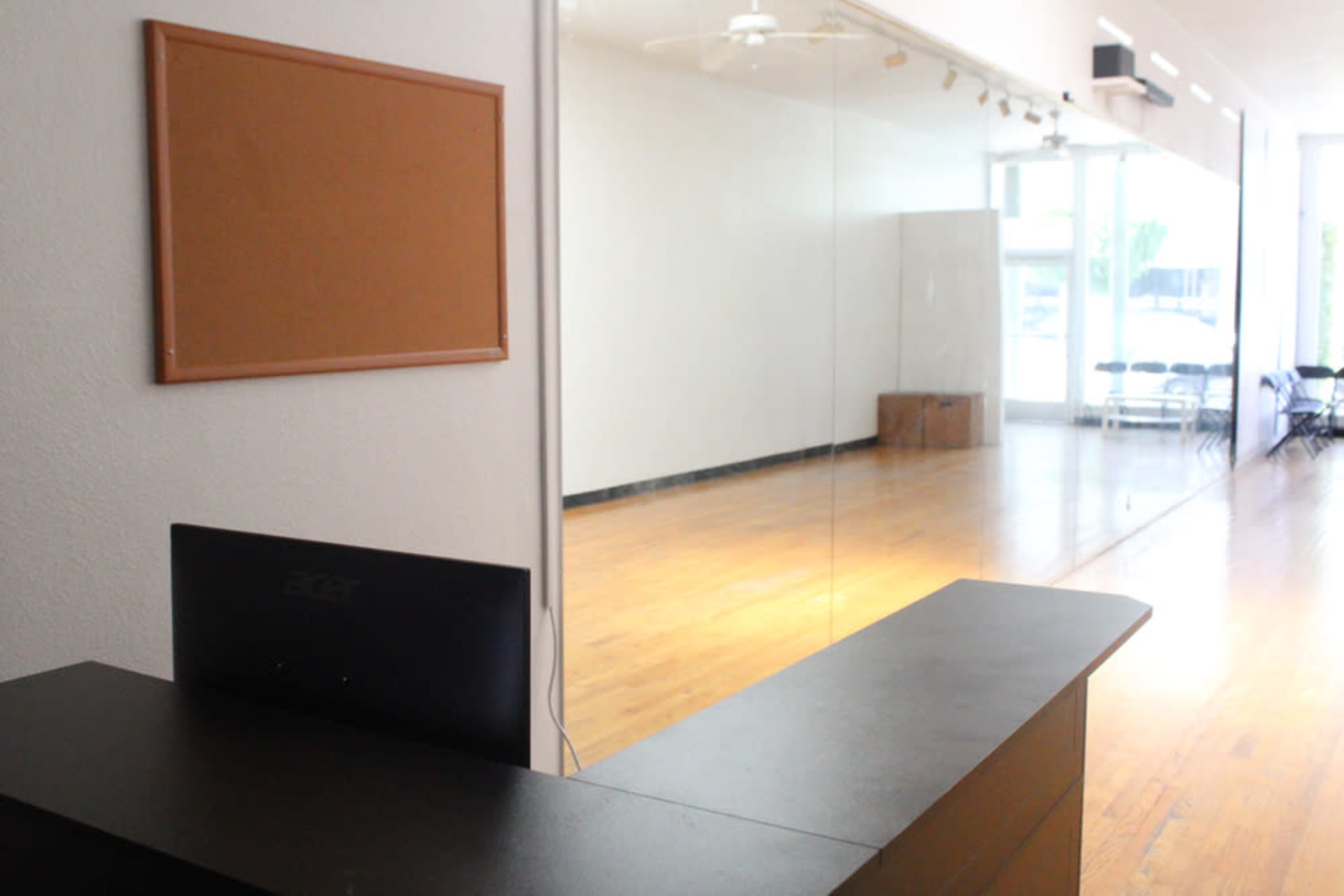The image shows a reception area with a dark countertop and a computer, leading into a large, empty room with a mirror and wooden flooring.