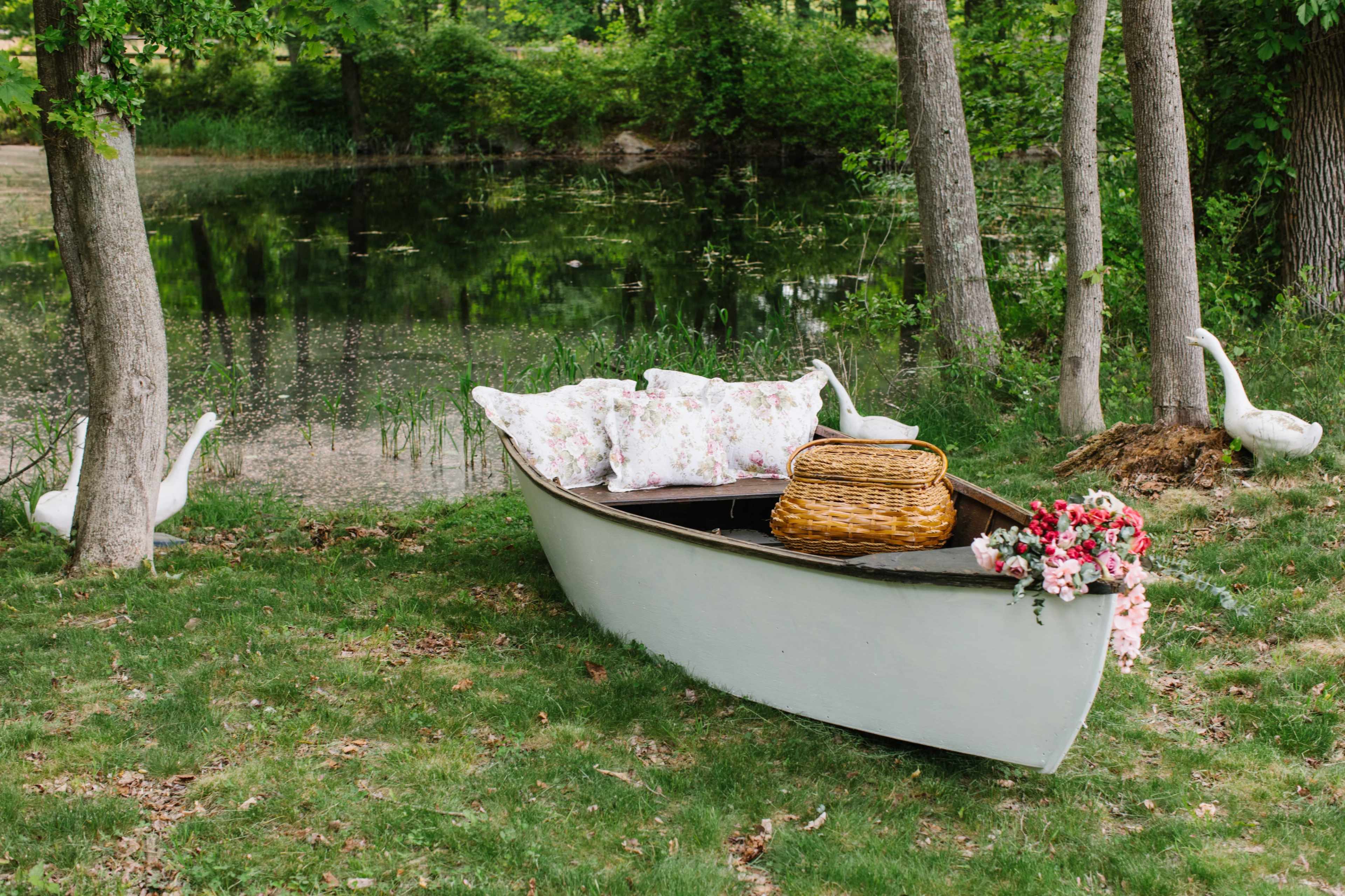 A canoe with floral cushions and a wicker basket is positioned near a pond, surrounded by trees and two white geese.