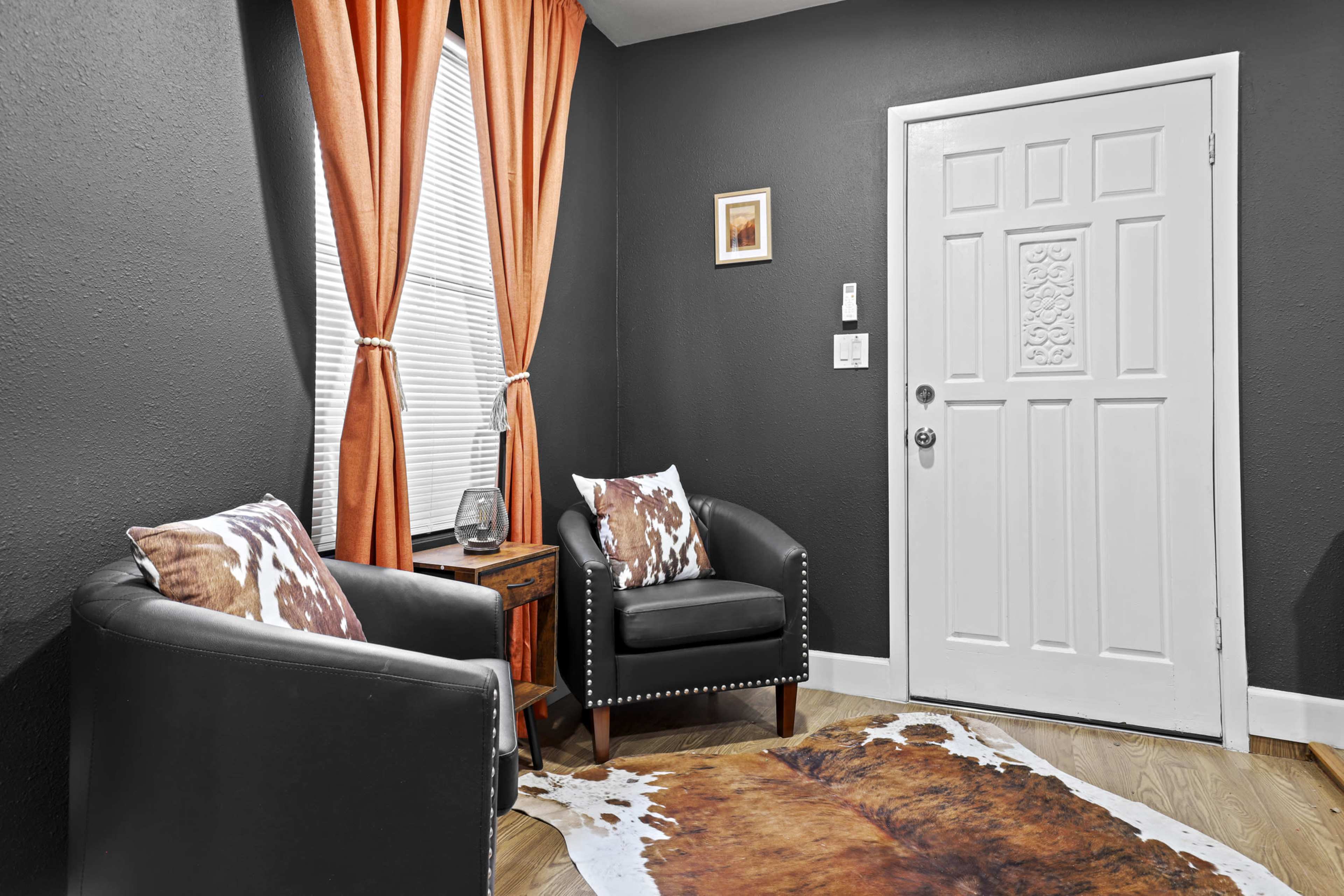 The image shows a small seating area with two black chairs, a wooden side table, a white door, and orange curtains against a dark wall.