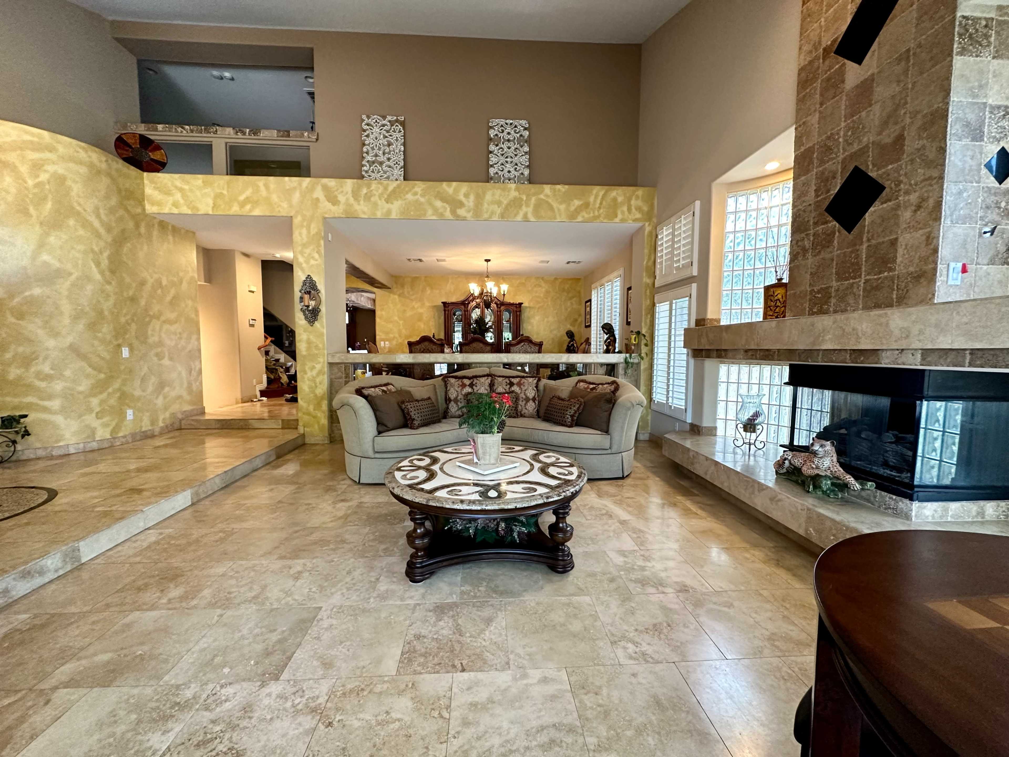 The image shows a spacious living room featuring a beige sofa, a round coffee table with a decorative surface, and a stone fireplace, with walls painted in a warm yellow tone.