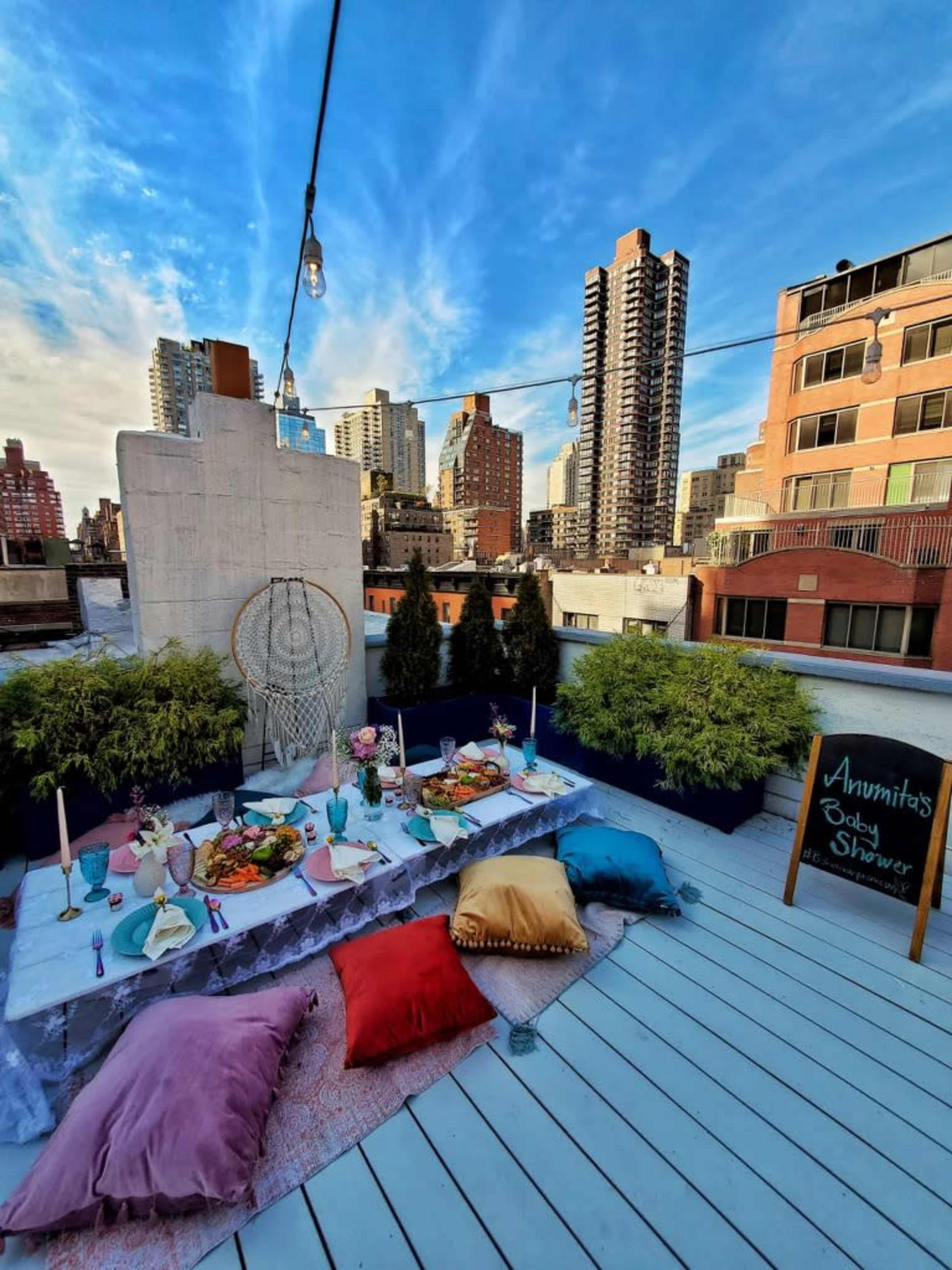 A rooftop garden features a vibrant picnic setup with a decorated table, cushions, and city skyline in the background.