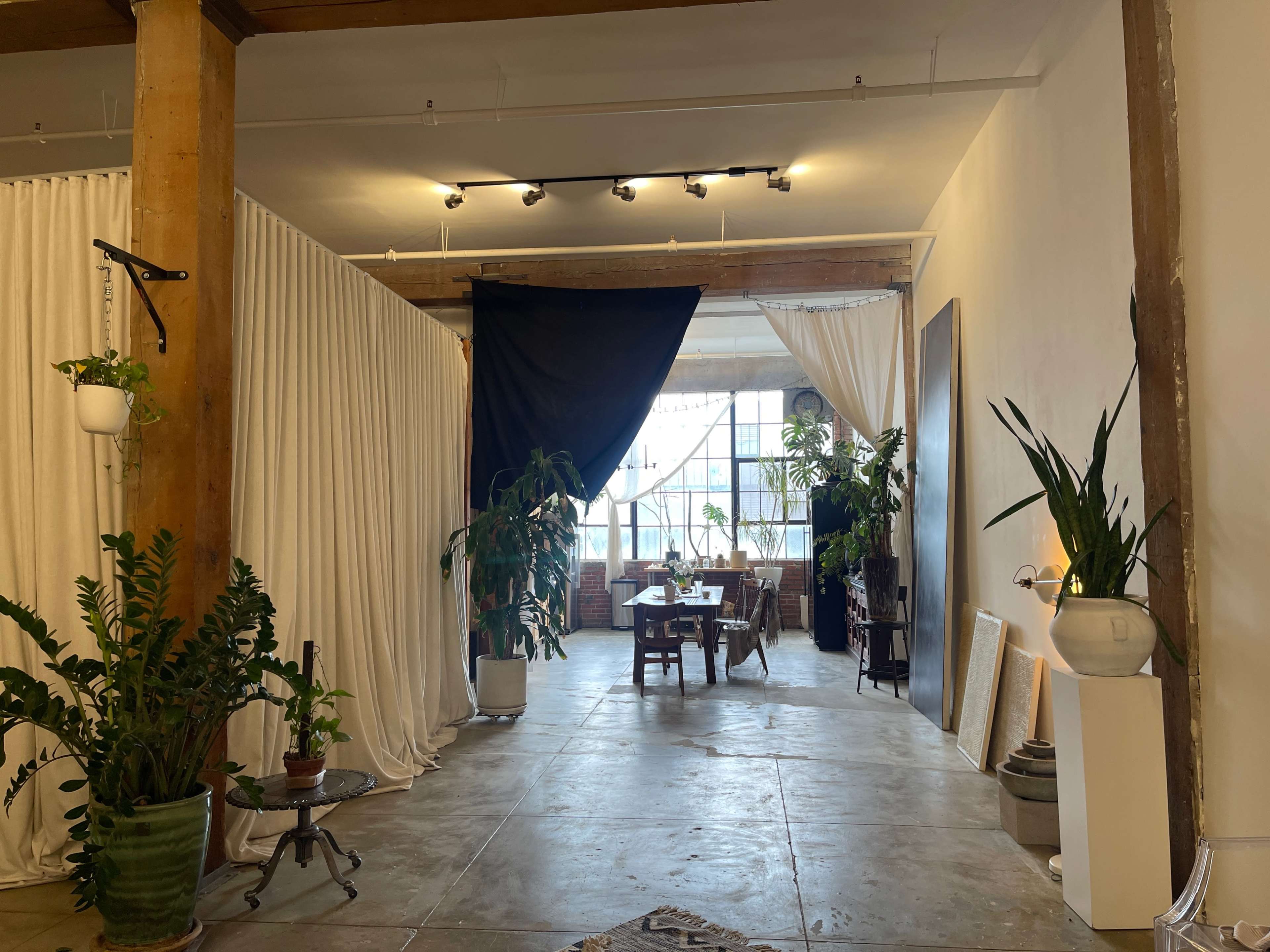 A spacious interior with exposed wooden beams, white curtains creating separate areas, and a table surrounded by plants and furniture.
