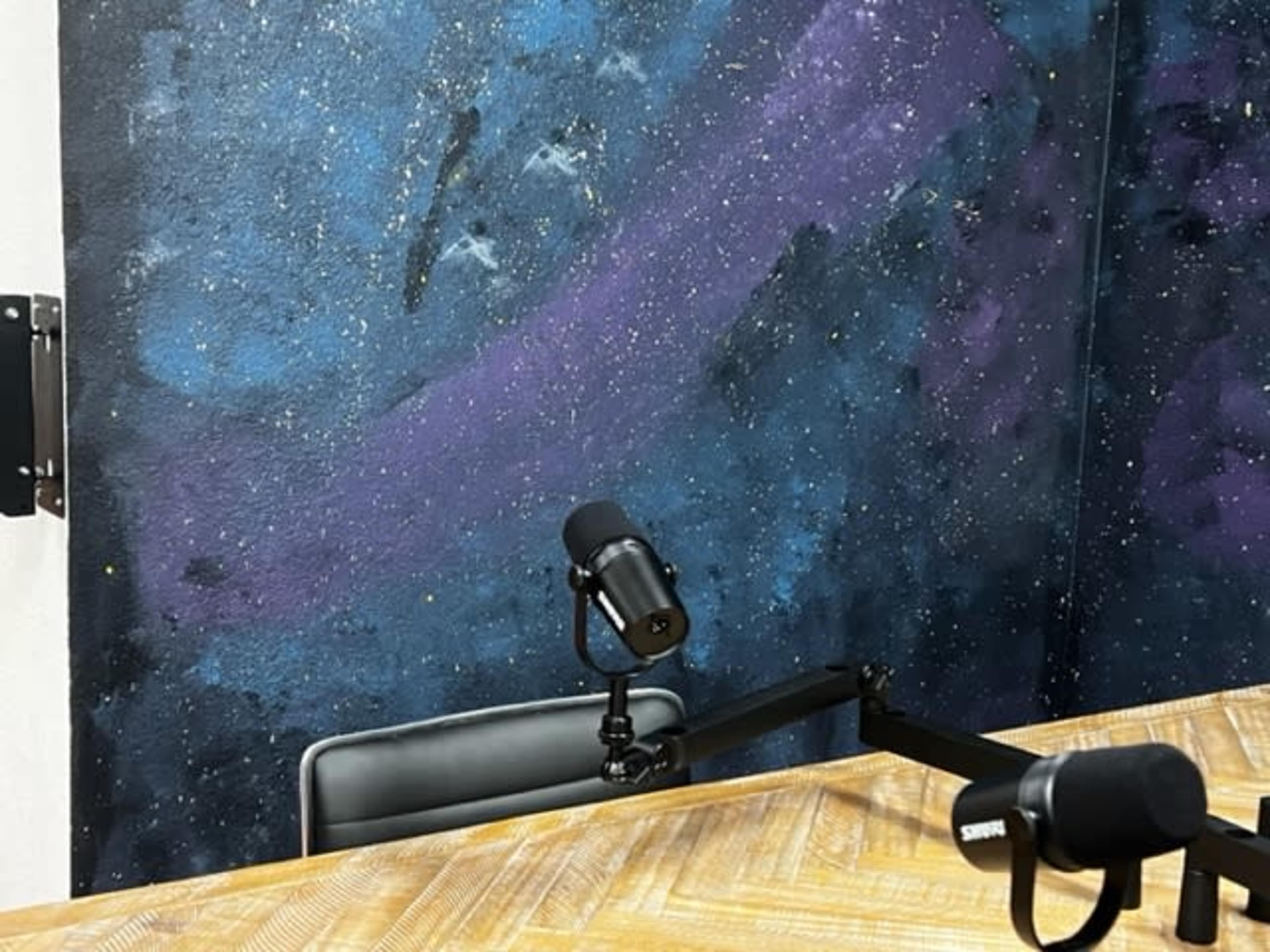The image shows a microphone and a chair positioned at a table in front of a colorful, painted wall resembling a starry night sky.