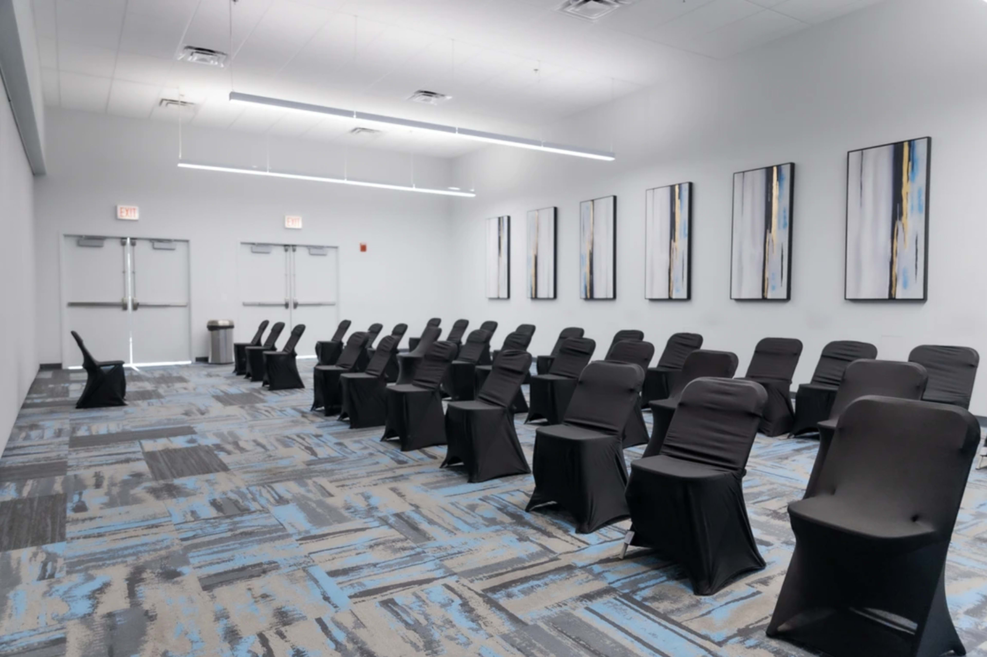 A row of black chairs covered with slipcovers is arranged facing a blank wall adorned with four abstract artworks in a well-lit room.