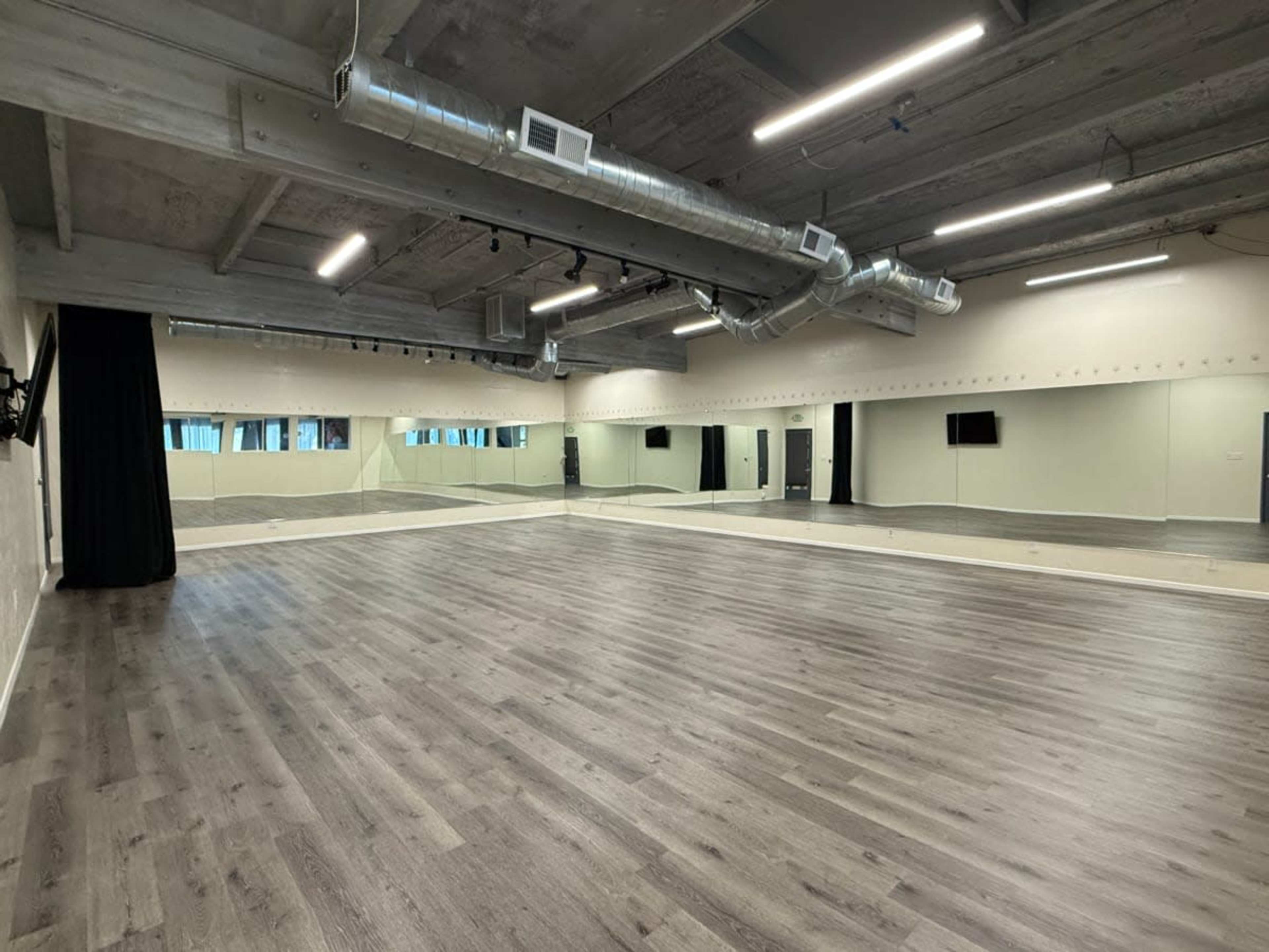 The image shows a spacious dance studio featuring hardwood flooring, large mirrors along one wall, and overhead lighting.