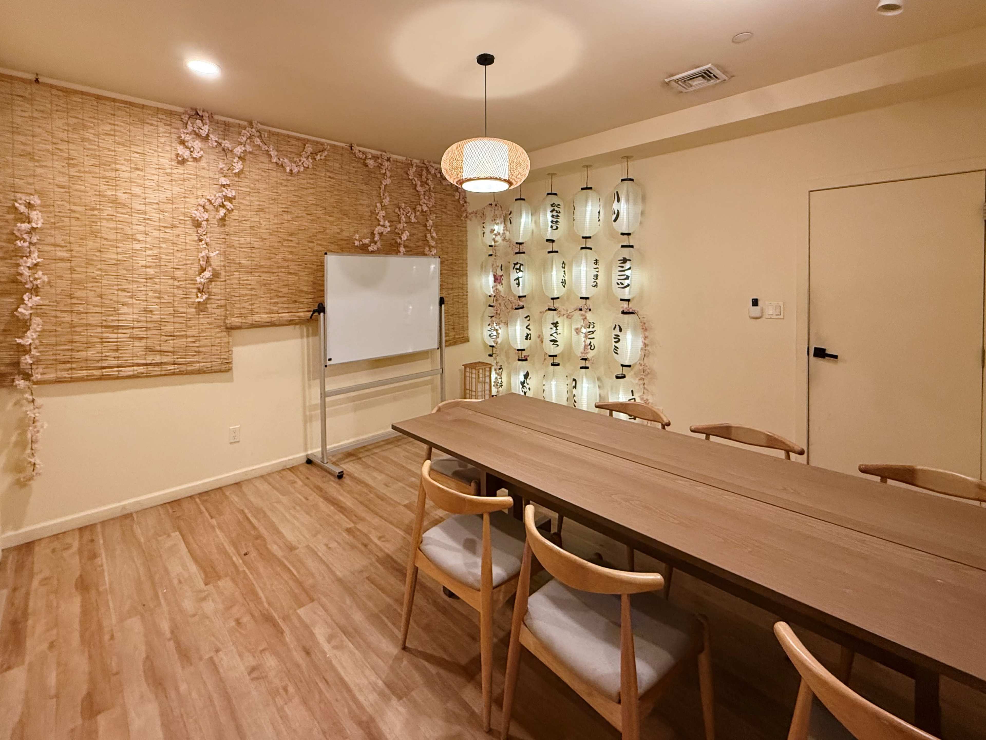 A small meeting room features a long wooden table surrounded by chairs, with a whiteboard on one side and decorative lanterns on the wall.