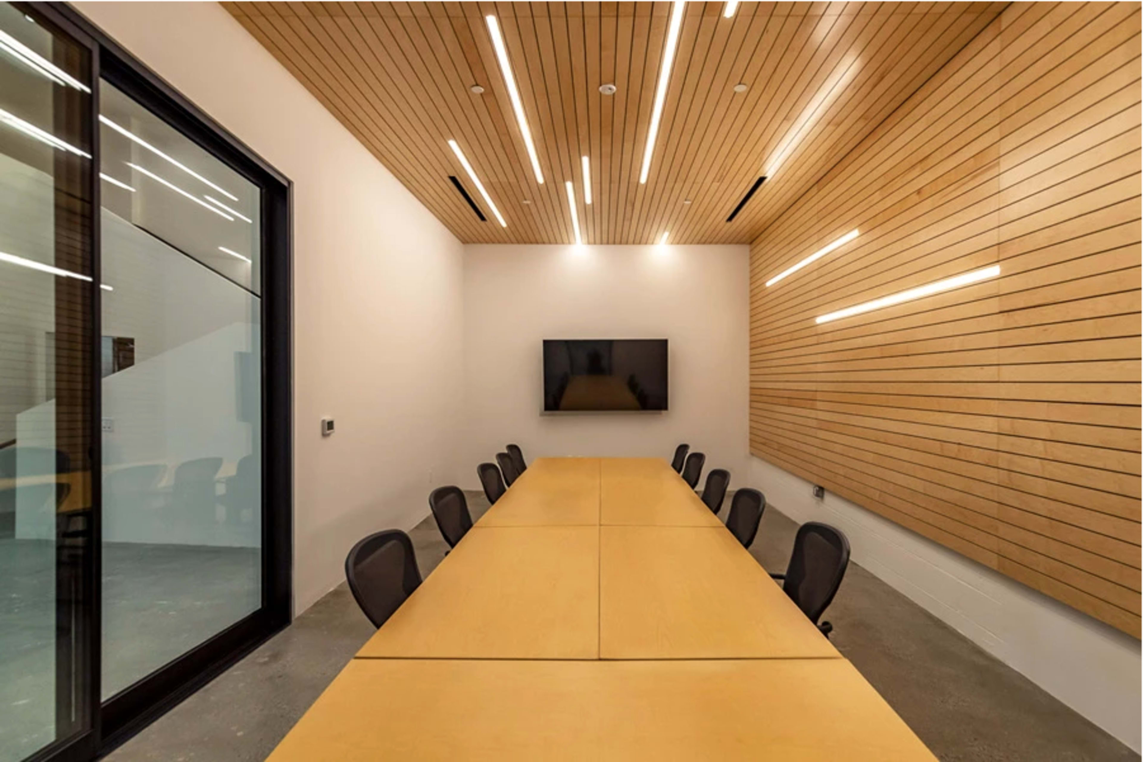 A modern conference room with a long wooden table, black chairs, and a large screen on the wall, illuminated by linear ceiling lights.
