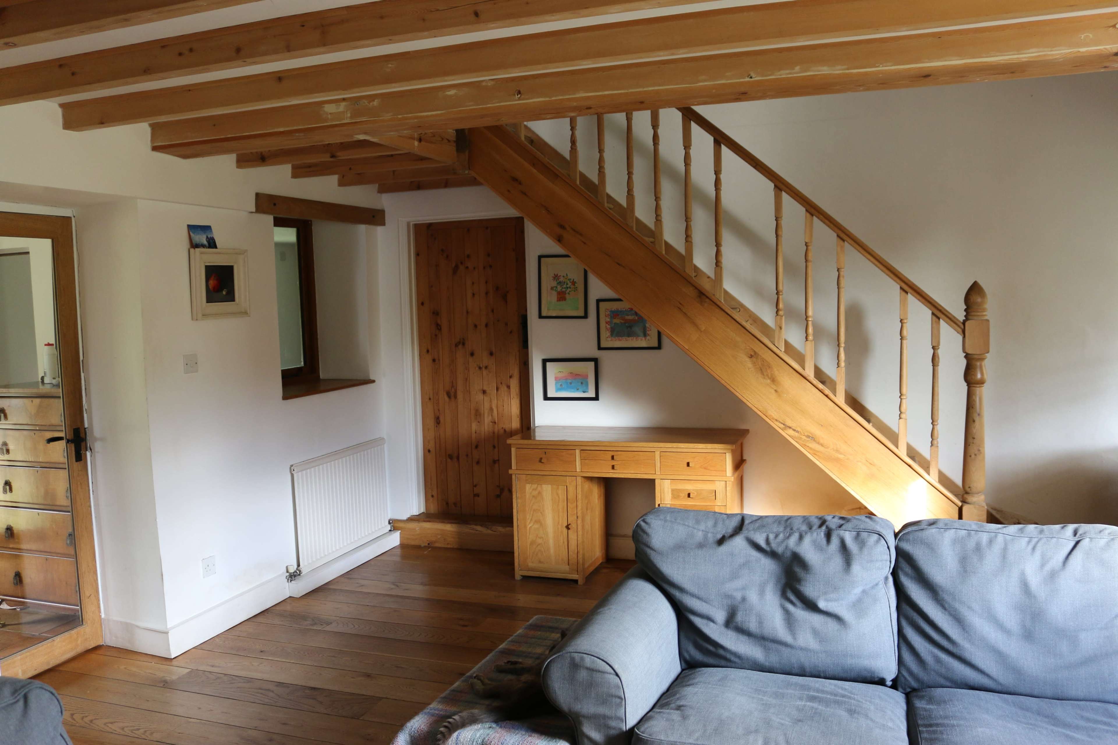 The image shows a living room featuring a wooden staircase, a desk, and a cozy couch, with framed artwork on the walls.