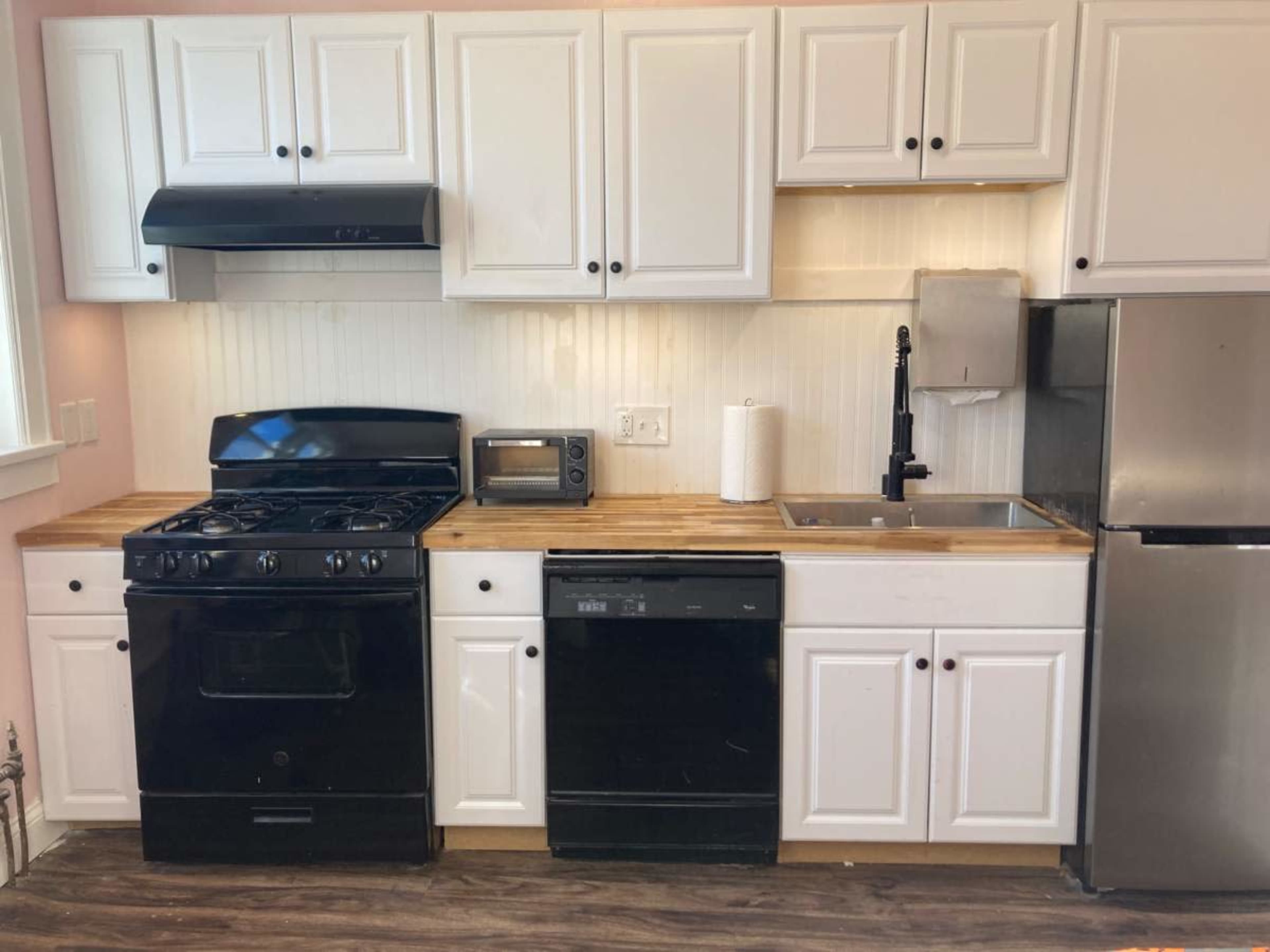 The image shows a compact kitchen featuring white cabinetry, a black stove, oven, microwave, dishwasher, stainless steel sink, and a fridge with a wooden countertop.