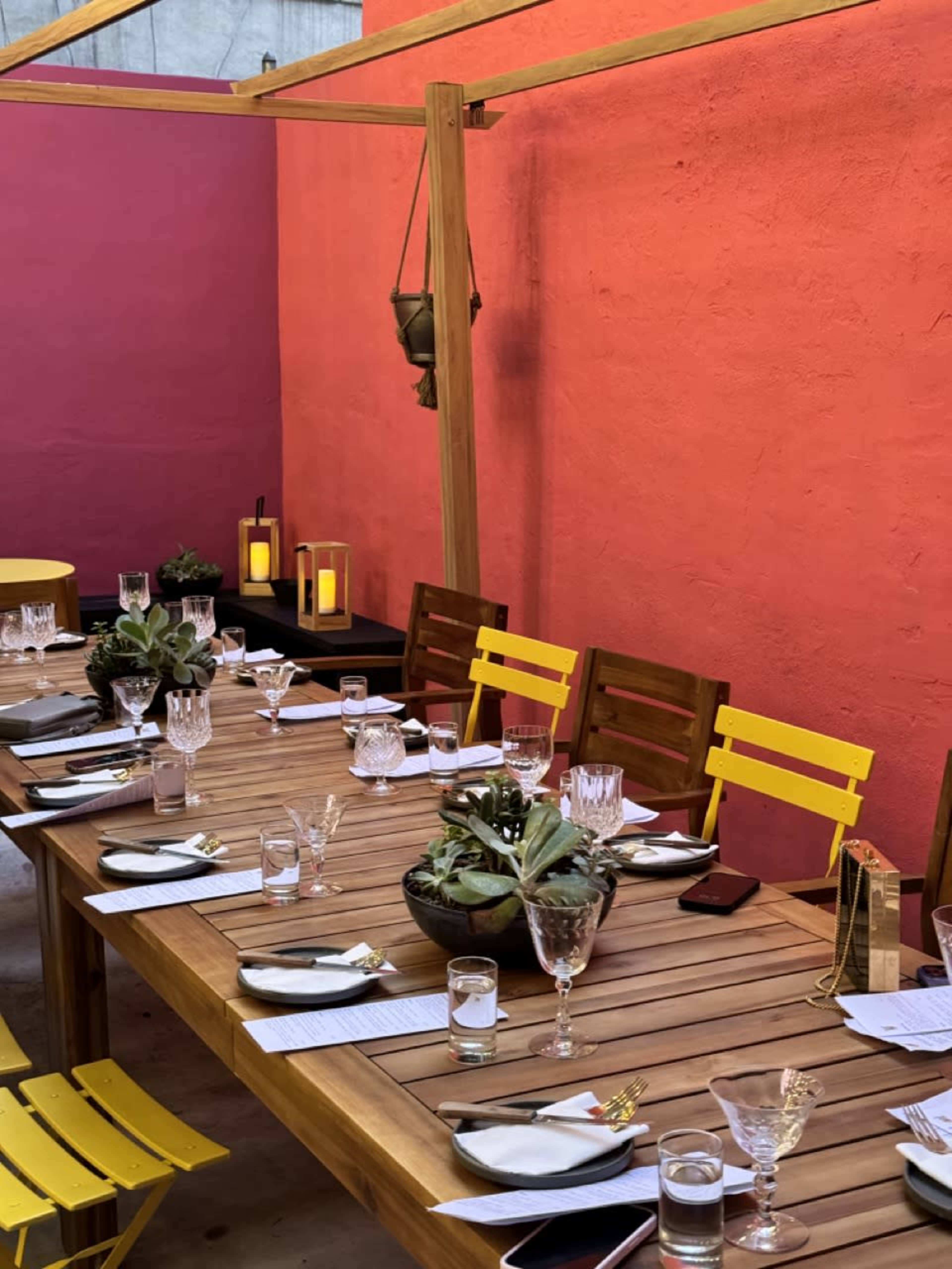 A long wooden table is set with glassware and utensils, accompanied by potted plants and lanterns, against a backdrop of vibrant pink walls.