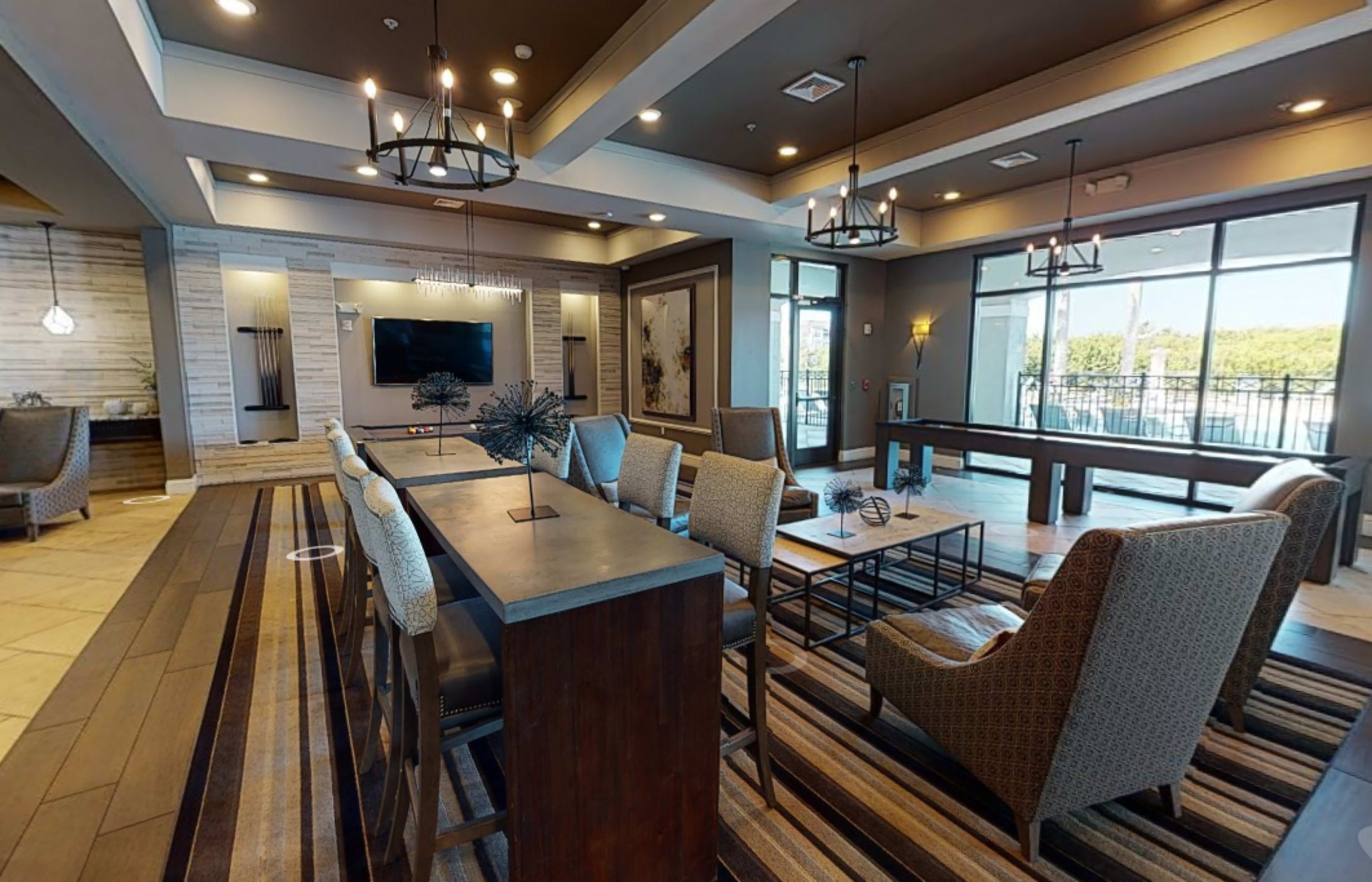 A modern lounge area with a table, several chairs, and large windows overlooking a patio space.