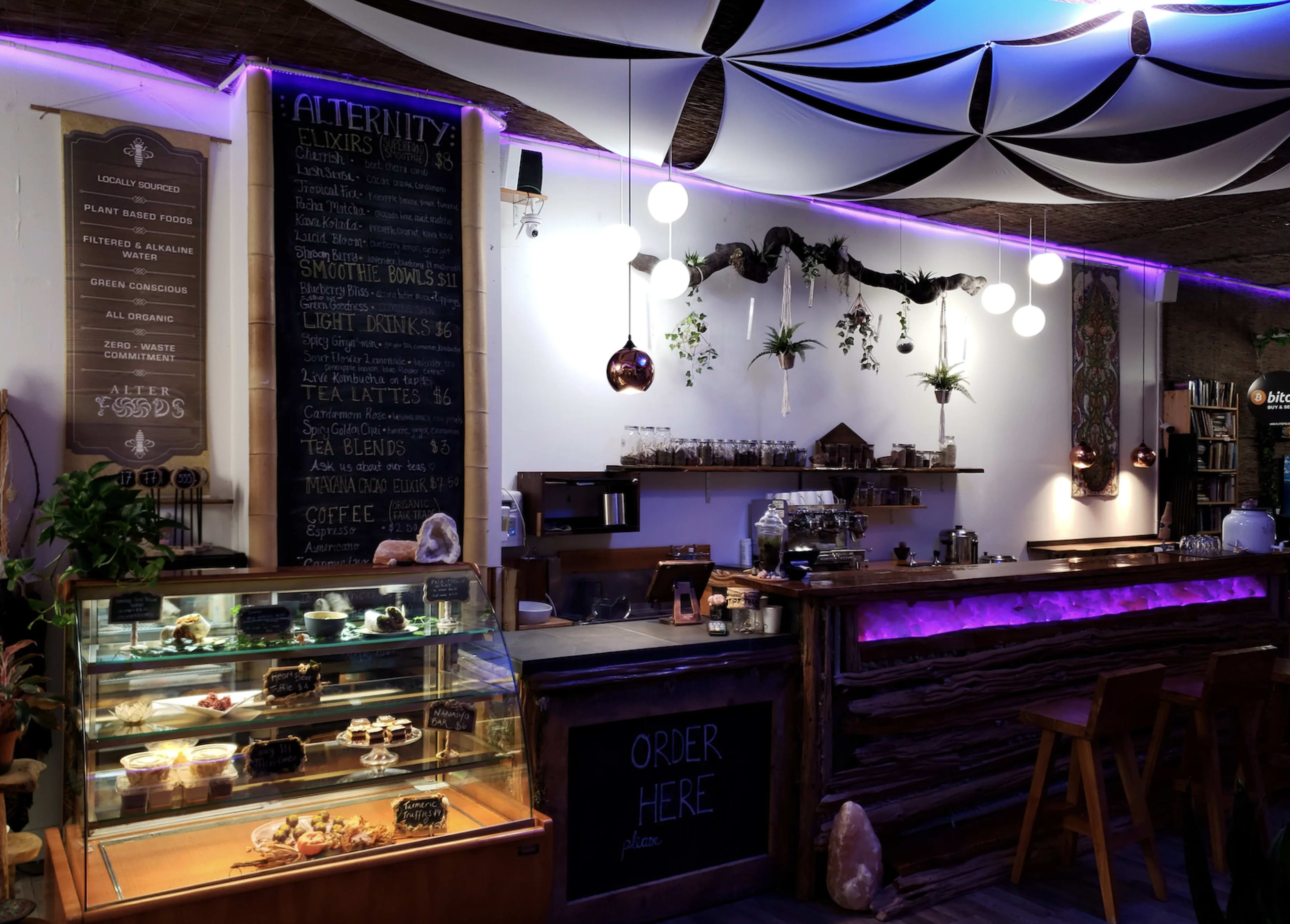 The image shows a cozy cafe interior with a wooden counter, a display case of desserts, and a menu board featuring elixirs and drinks under soft lighting and decorative plants.