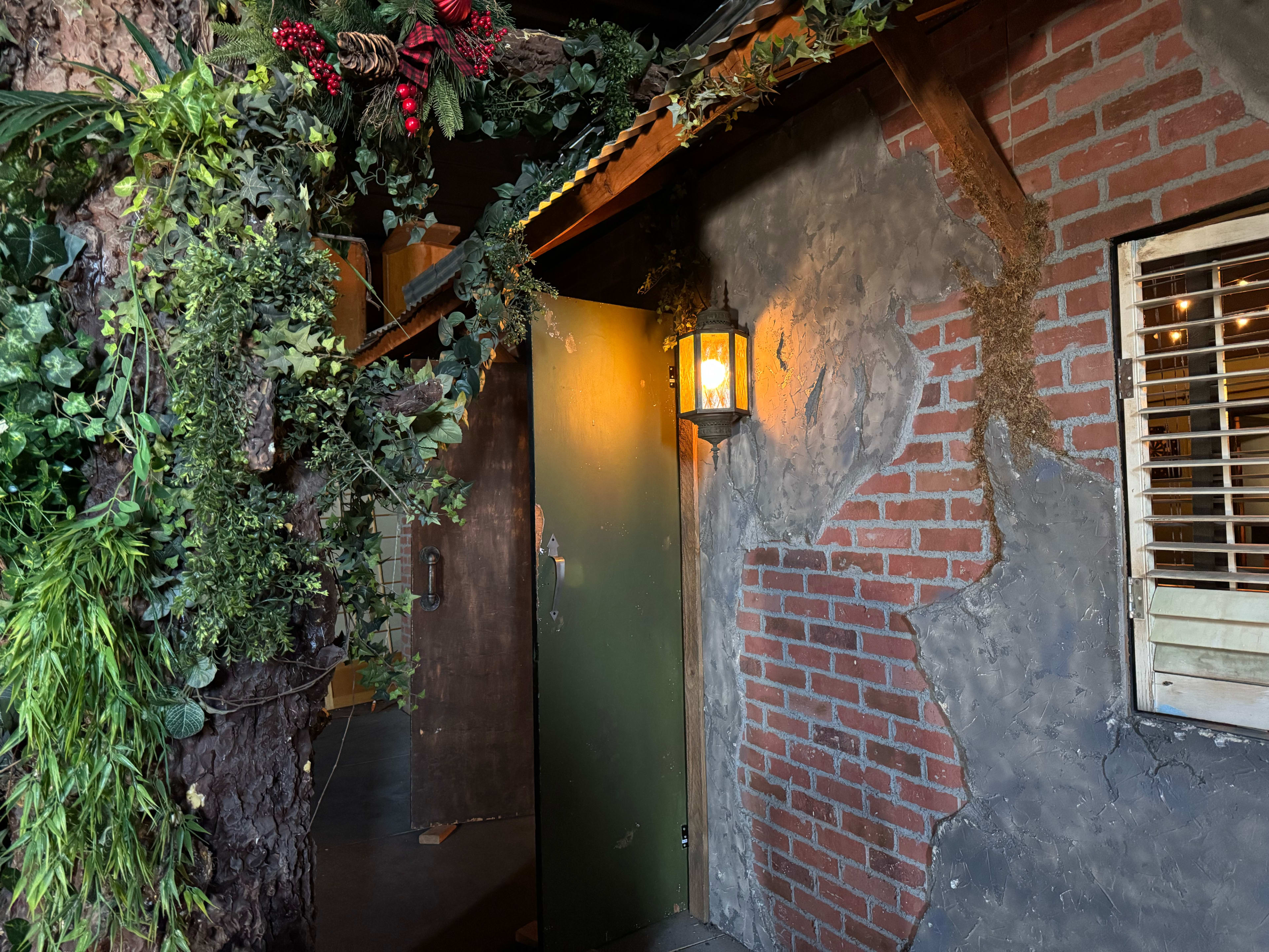 A weathered wooden door is framed by brick walls and greenery, with a lantern illuminating the entrance.