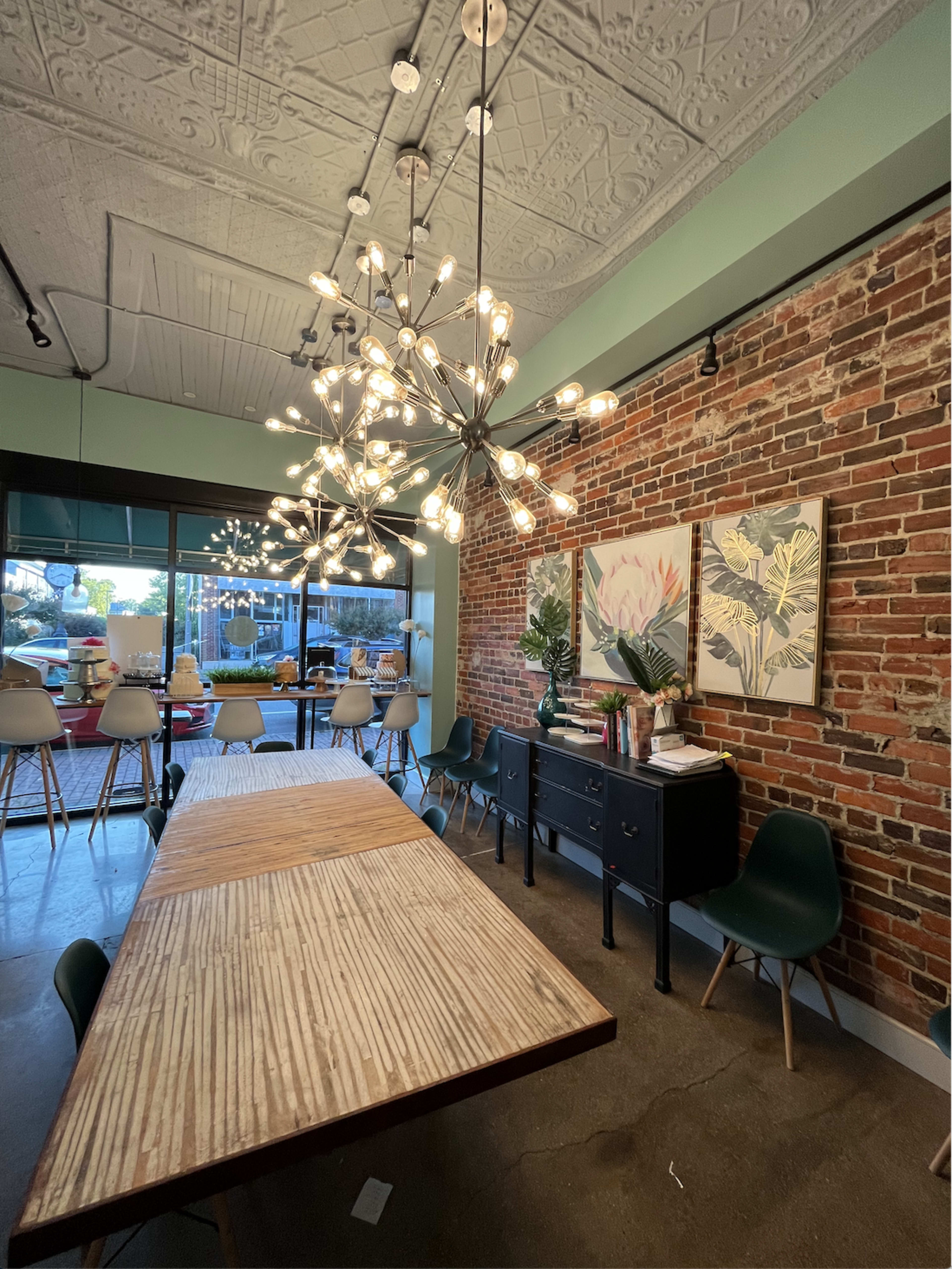 A dining area with a large wooden table, modern seating, a decorative cabinet, and two pieces of framed artwork beneath two contemporary light fixtures, all set against a brick wall.