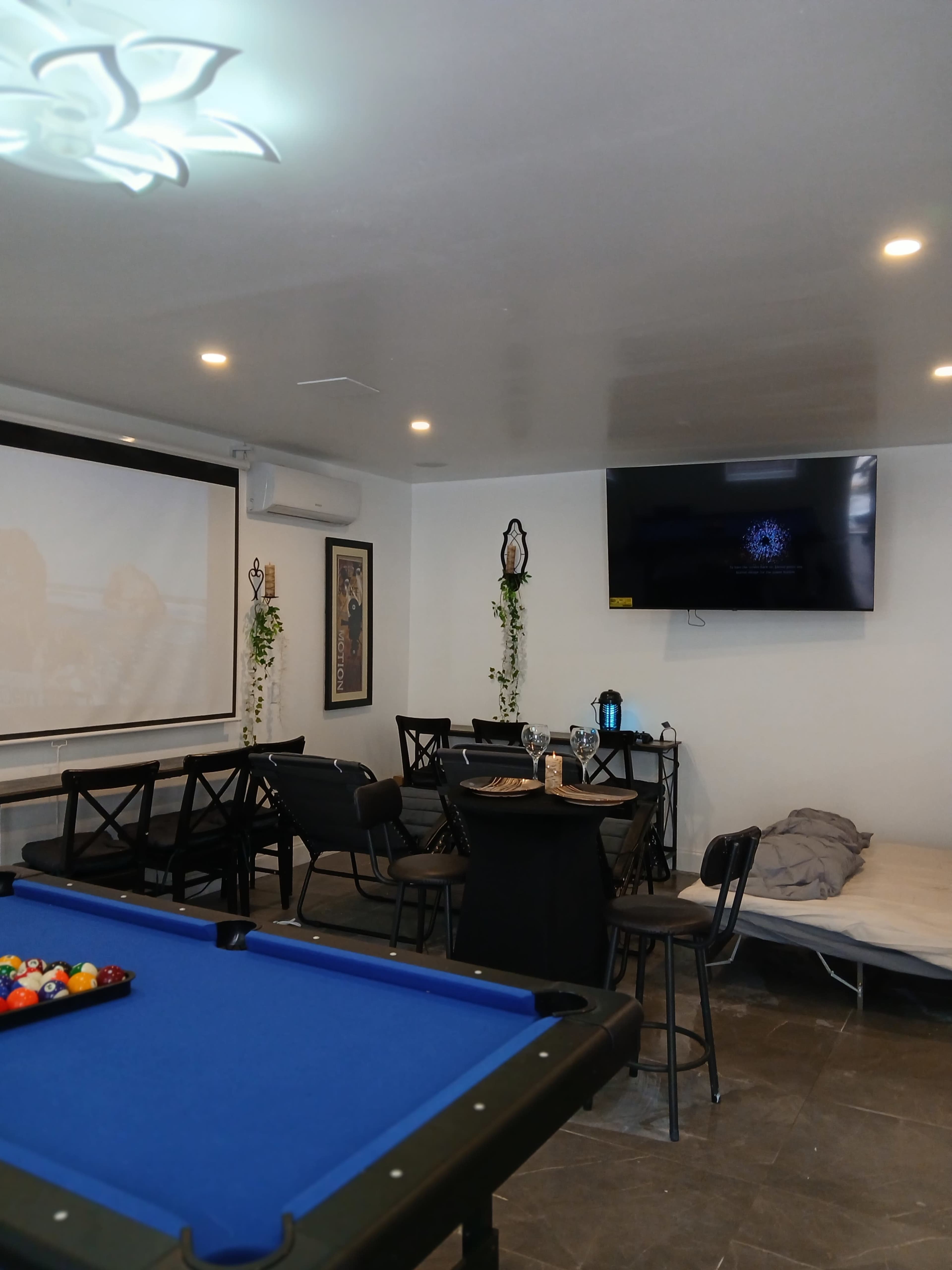 The image shows a room with a blue pool table, a dining area with chairs and a table, a large screen on the wall, and a couch on the floor.