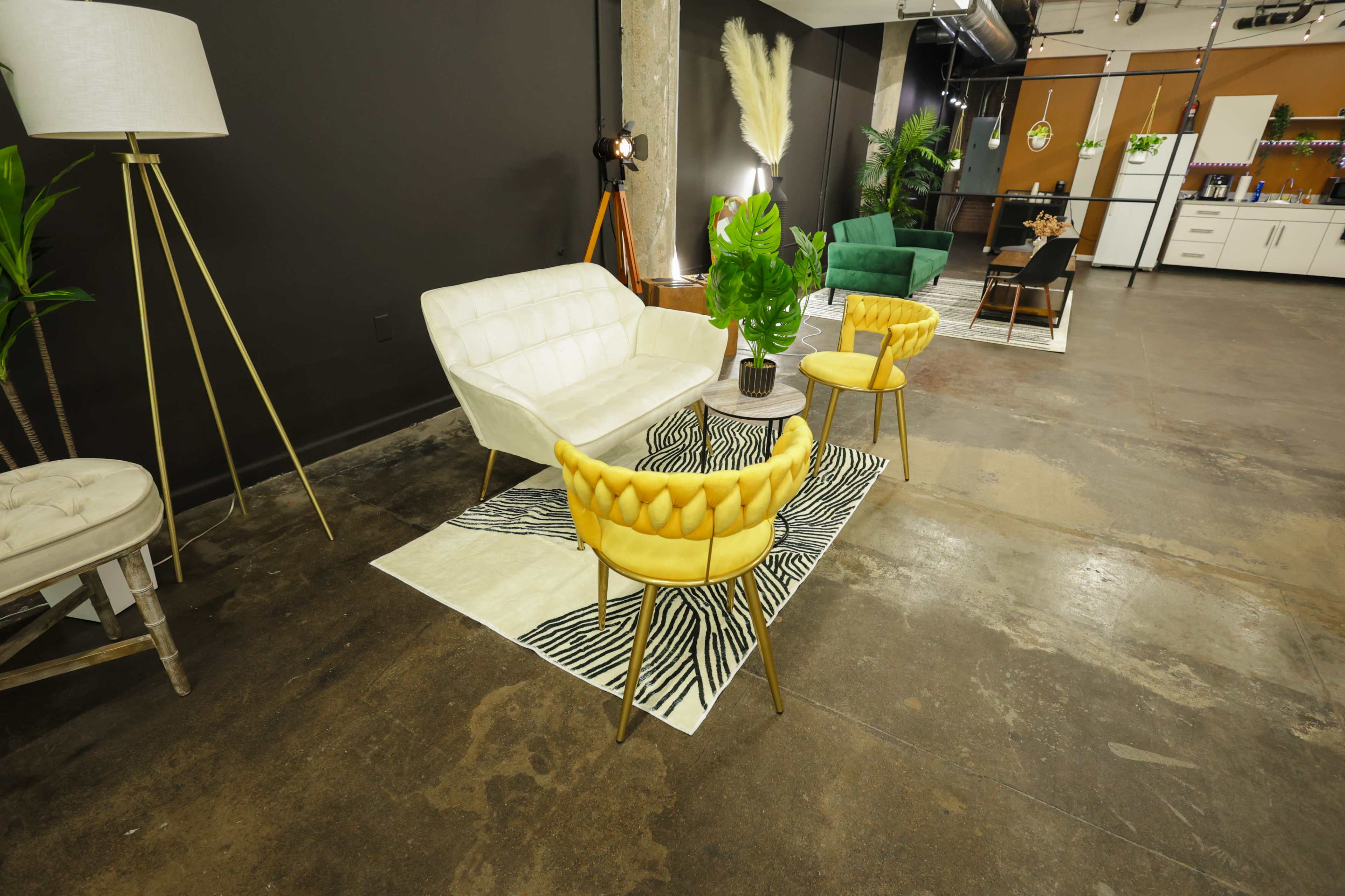 The image shows a stylish interior space featuring a white couch, two yellow chairs, and a small table on a patterned rug, with greenery and lighting equipment in the background.