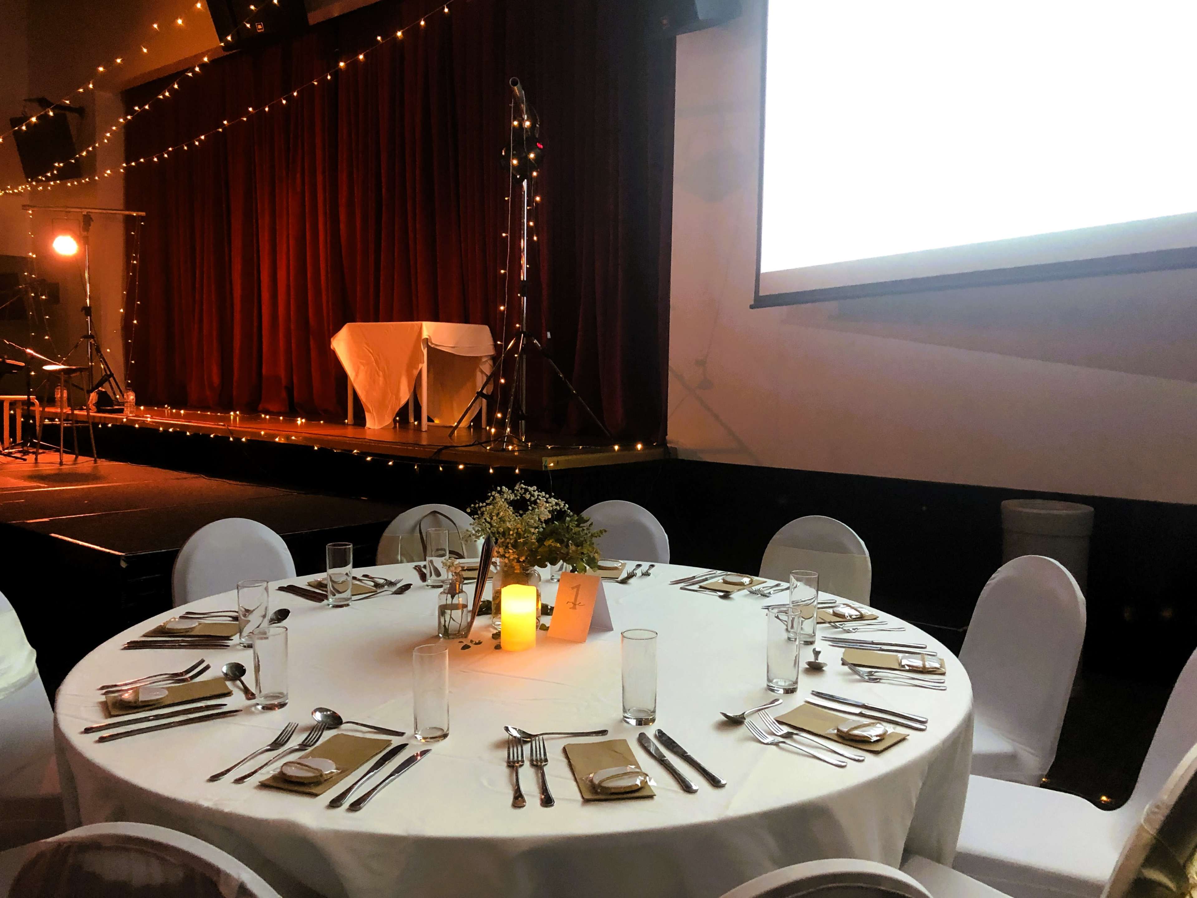 A round dining table is set with cutlery and glasses, surrounded by white chairs, in front of a stage with draped curtains and decorative lights.
