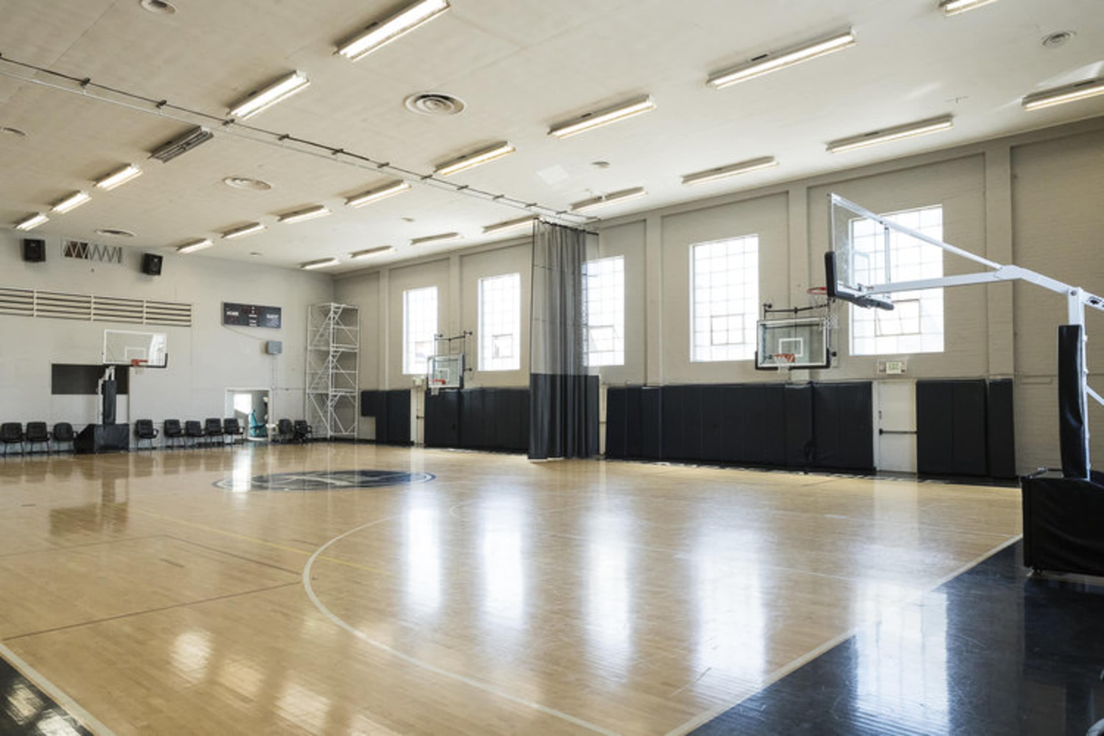 A spacious indoor basketball court with two hoops, wooden flooring, and large windows providing natural light.