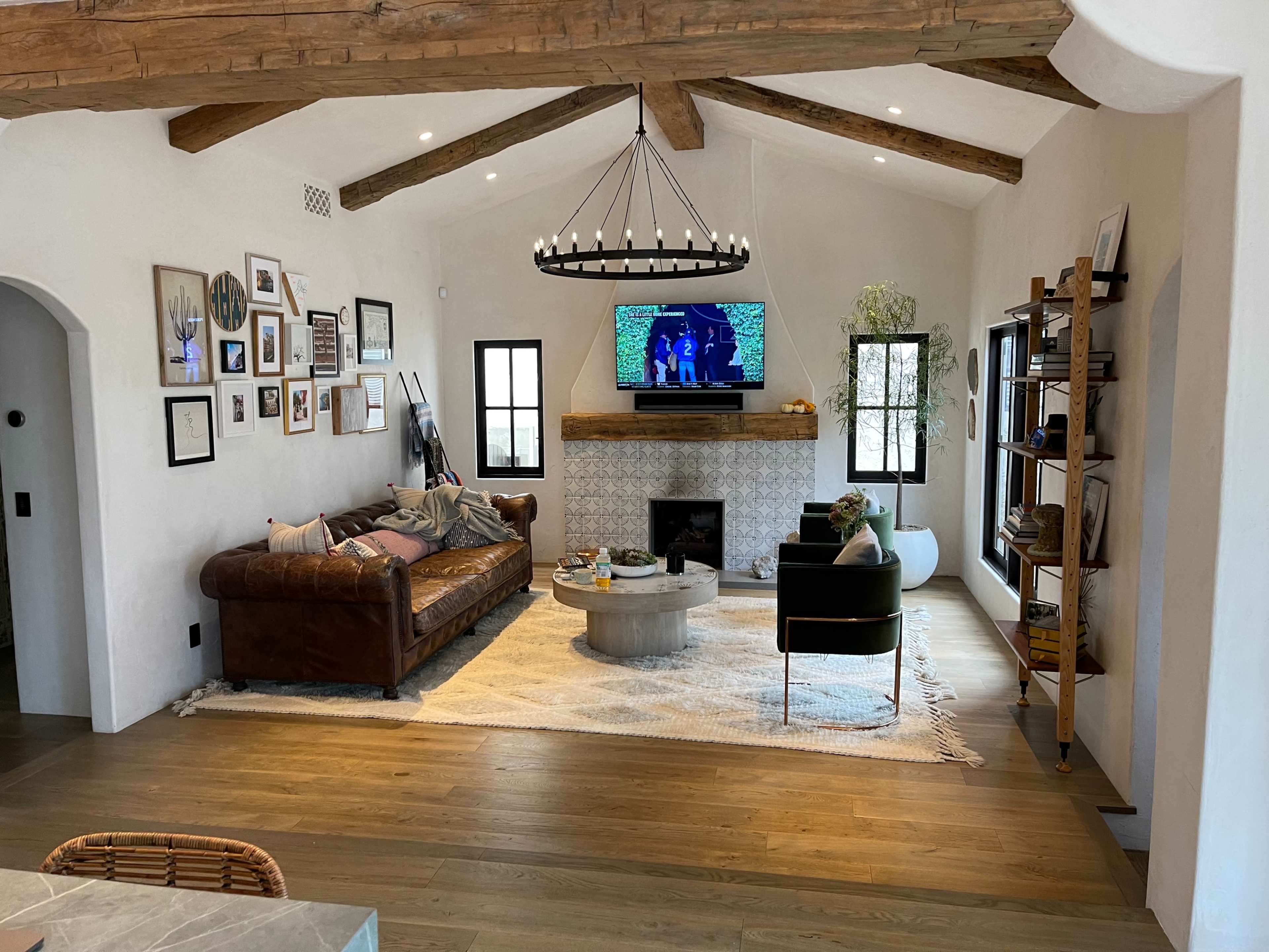 A cozy living room features a leather sofa, a round coffee table, and a television above a stone fireplace, all set beneath exposed wooden beams.