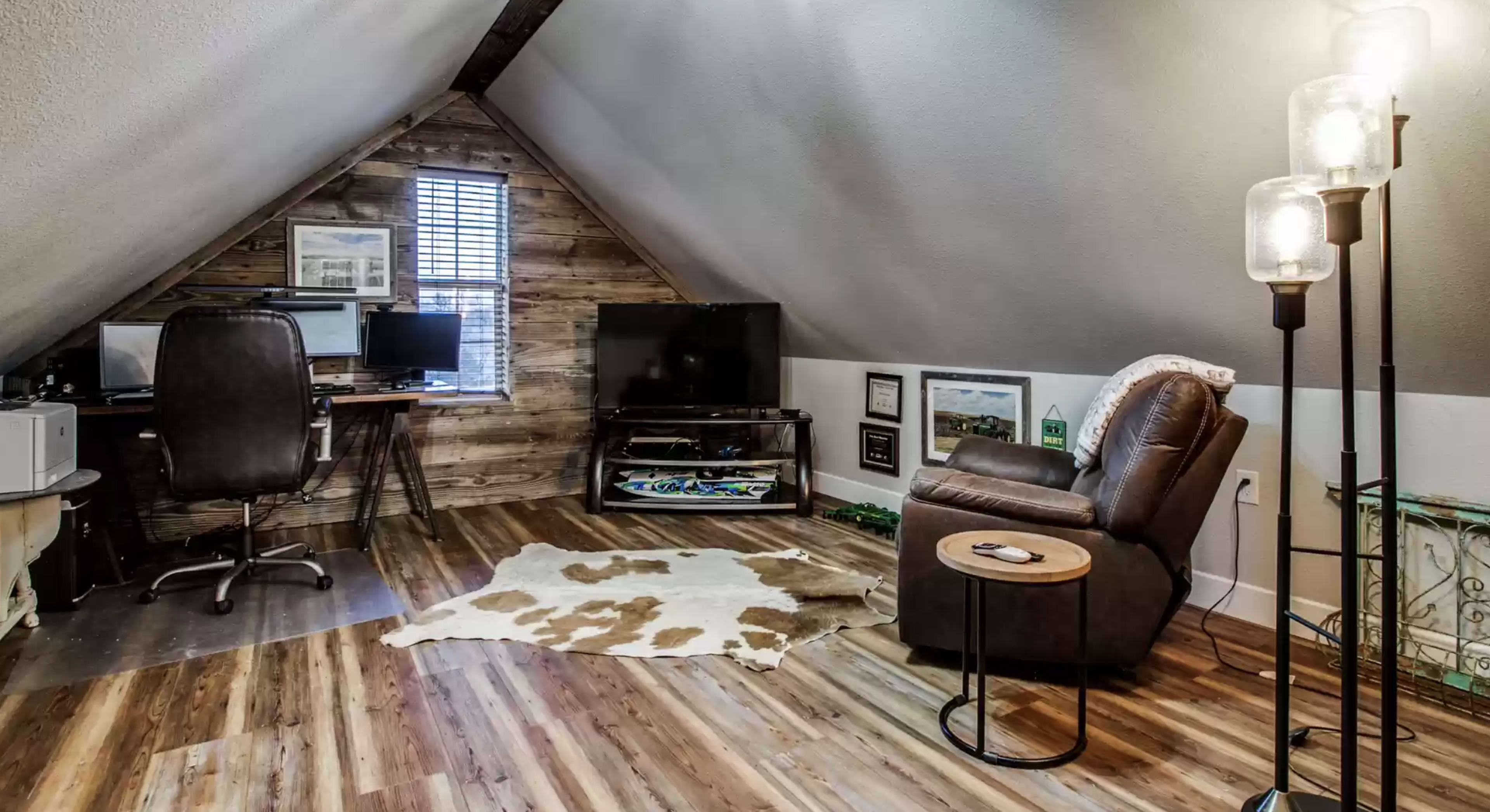 A cozy attic space with wooden walls, a recliner, a small side table, a flat-screen TV, and two workstations.