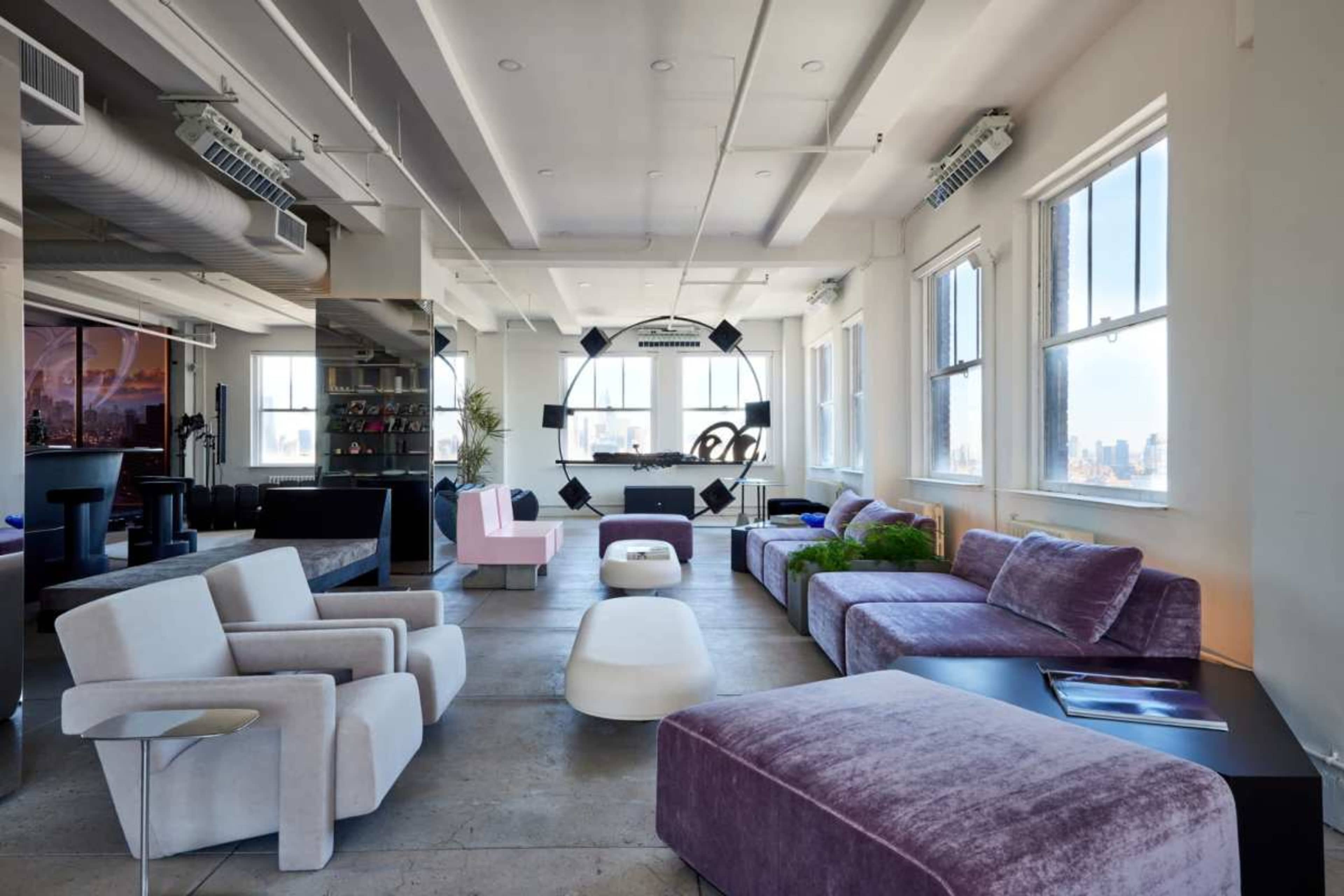 The image shows a modern, open-concept living space furnished with various colorful couches, a central table, and large windows providing natural light.