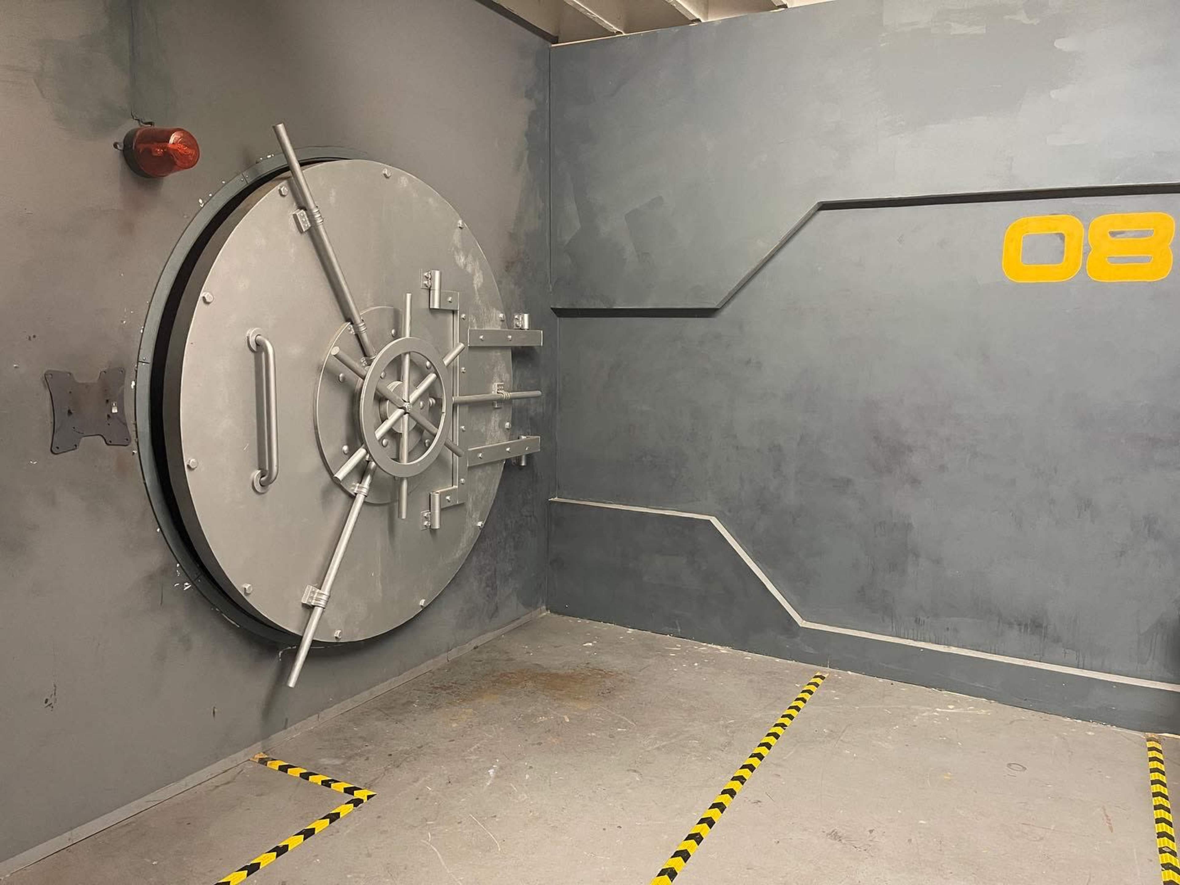 A large metallic vault door set into a gray wall, with a red light above and yellow markings on the floor.
