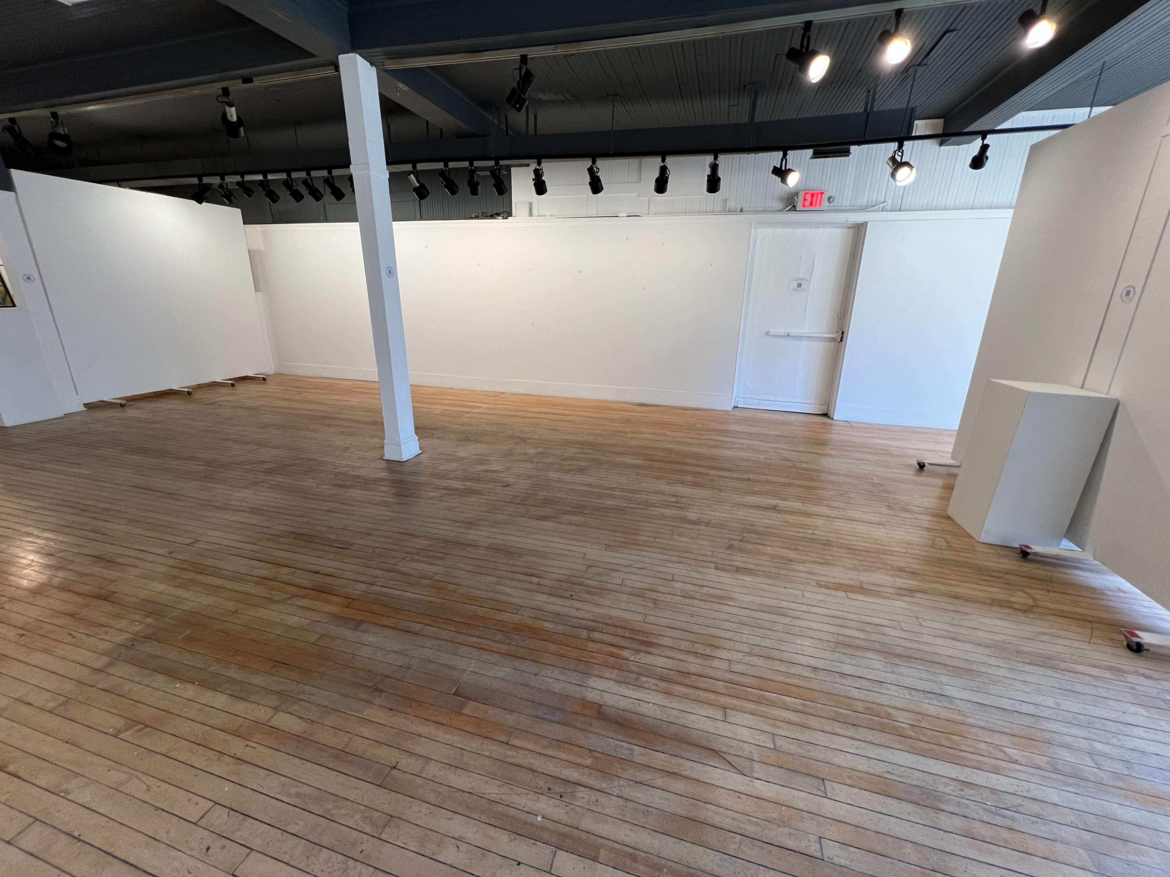 A spacious, empty room with wooden flooring and white walls, illuminated by overhead track lighting.