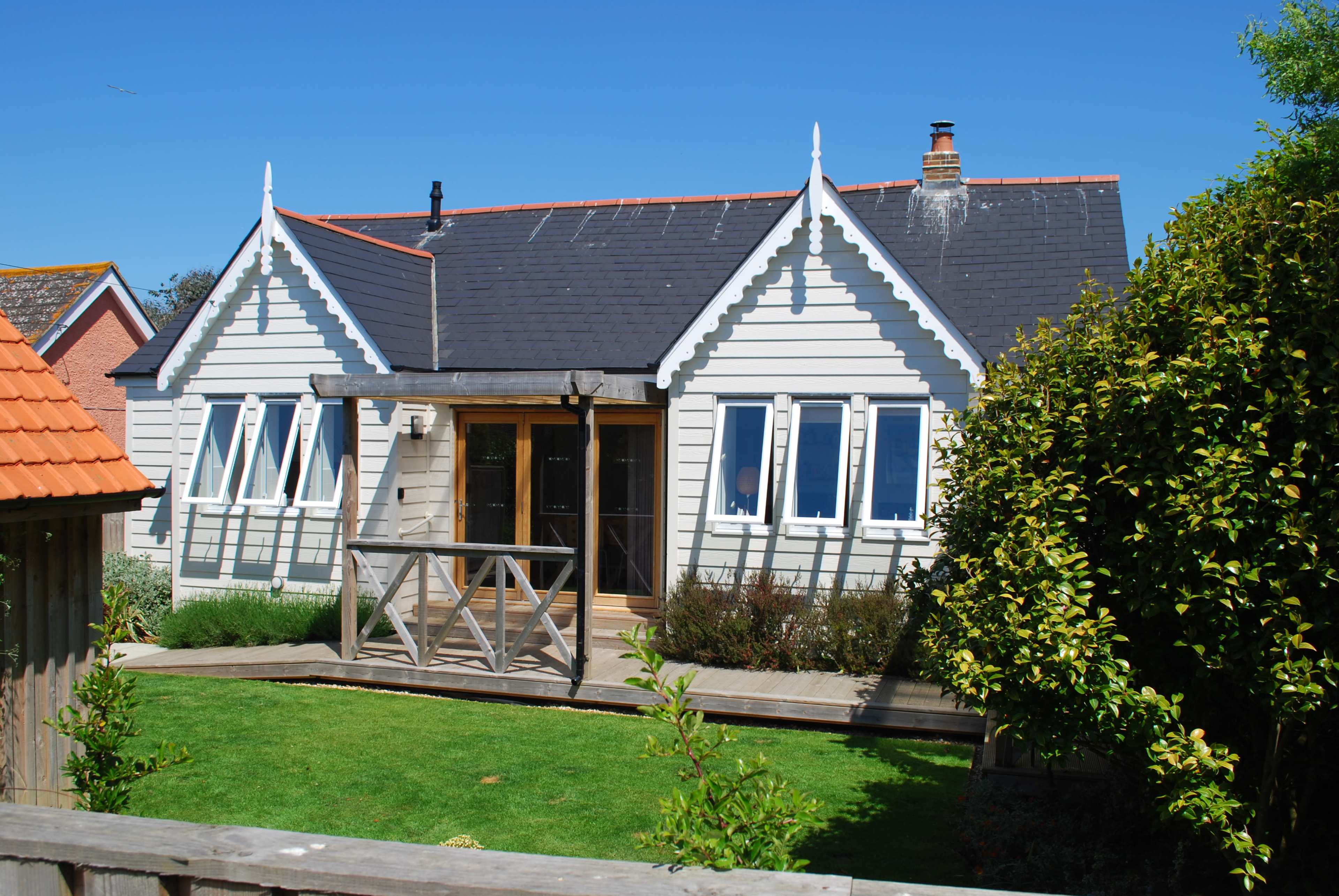 The image shows a light-colored wooden house with a gabled roof, large windows, and a front porch, surrounded by grass and shrubs.