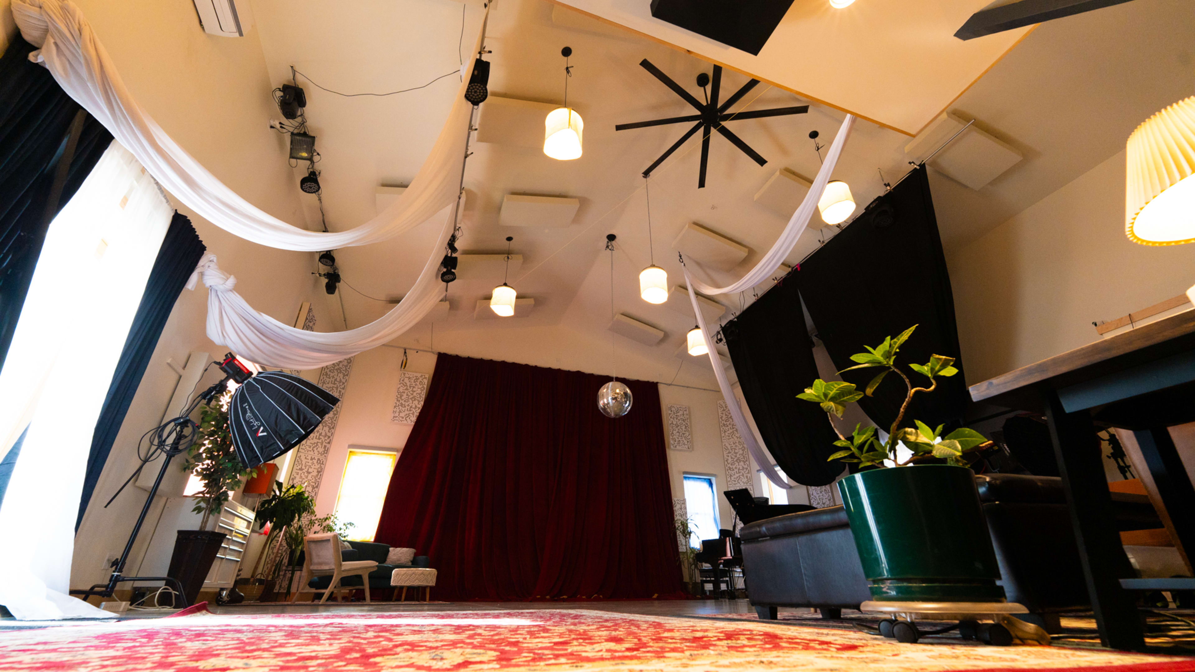 The image shows an interior space with high ceilings, draped fabric, a large red curtain at the back, and various plants and furniture arrangements.