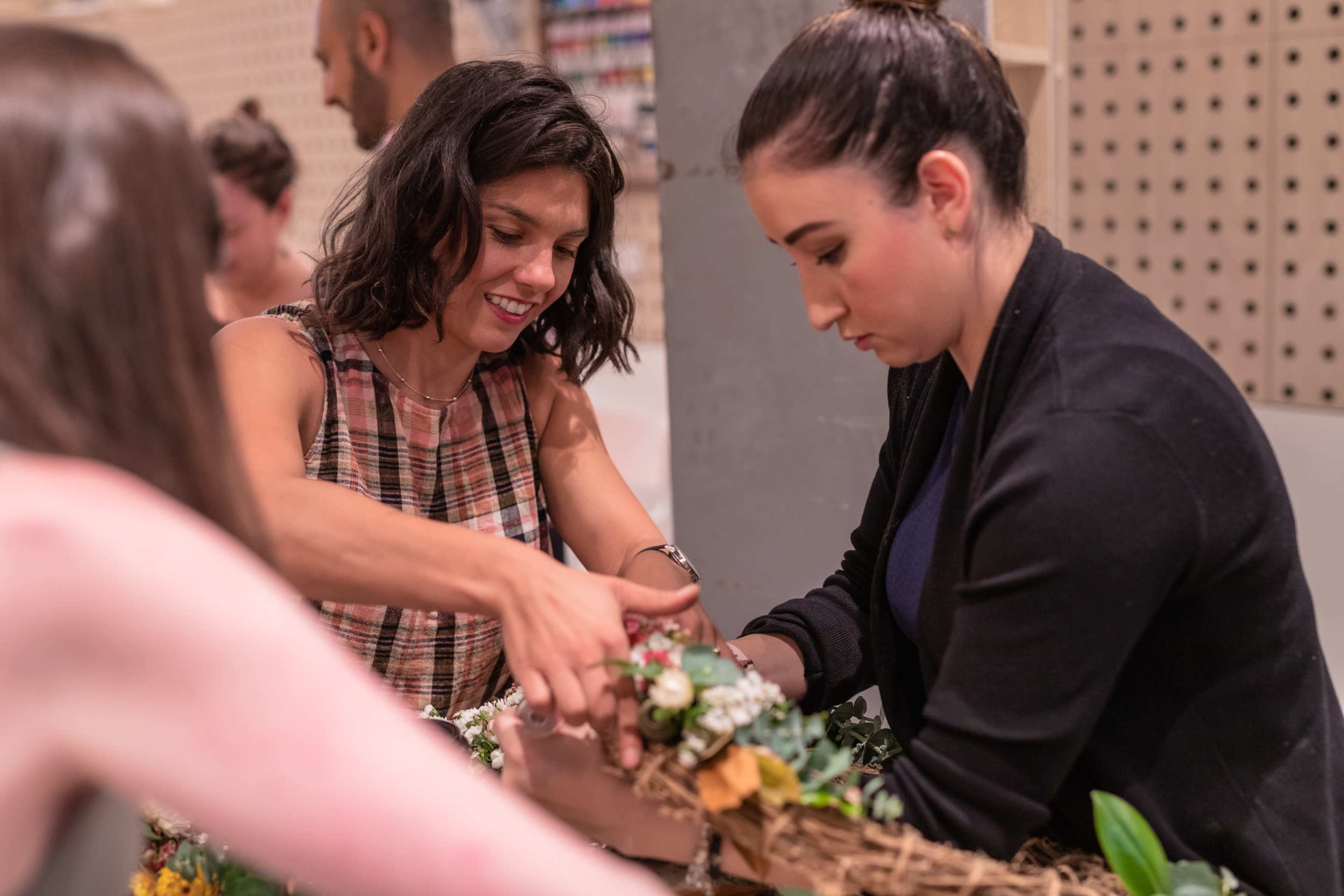 Two women collaborate on arranging flowers and greenery in a basket during a crafting session.