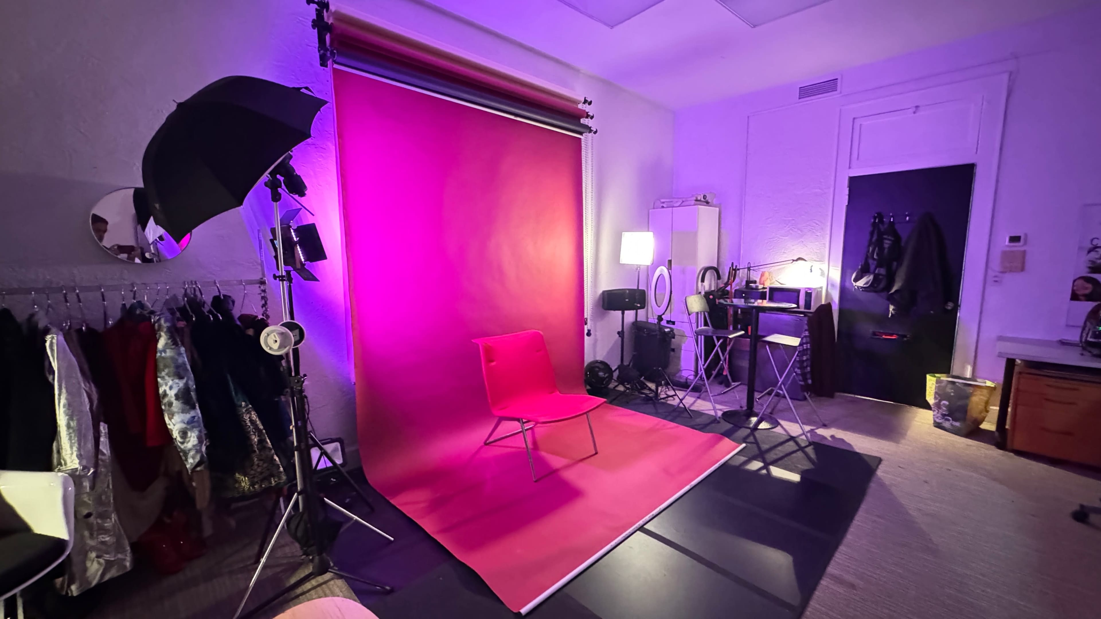 The image shows a photography studio setup with a pink backdrop, a chair in the center, lighting equipment, and various props along the side.