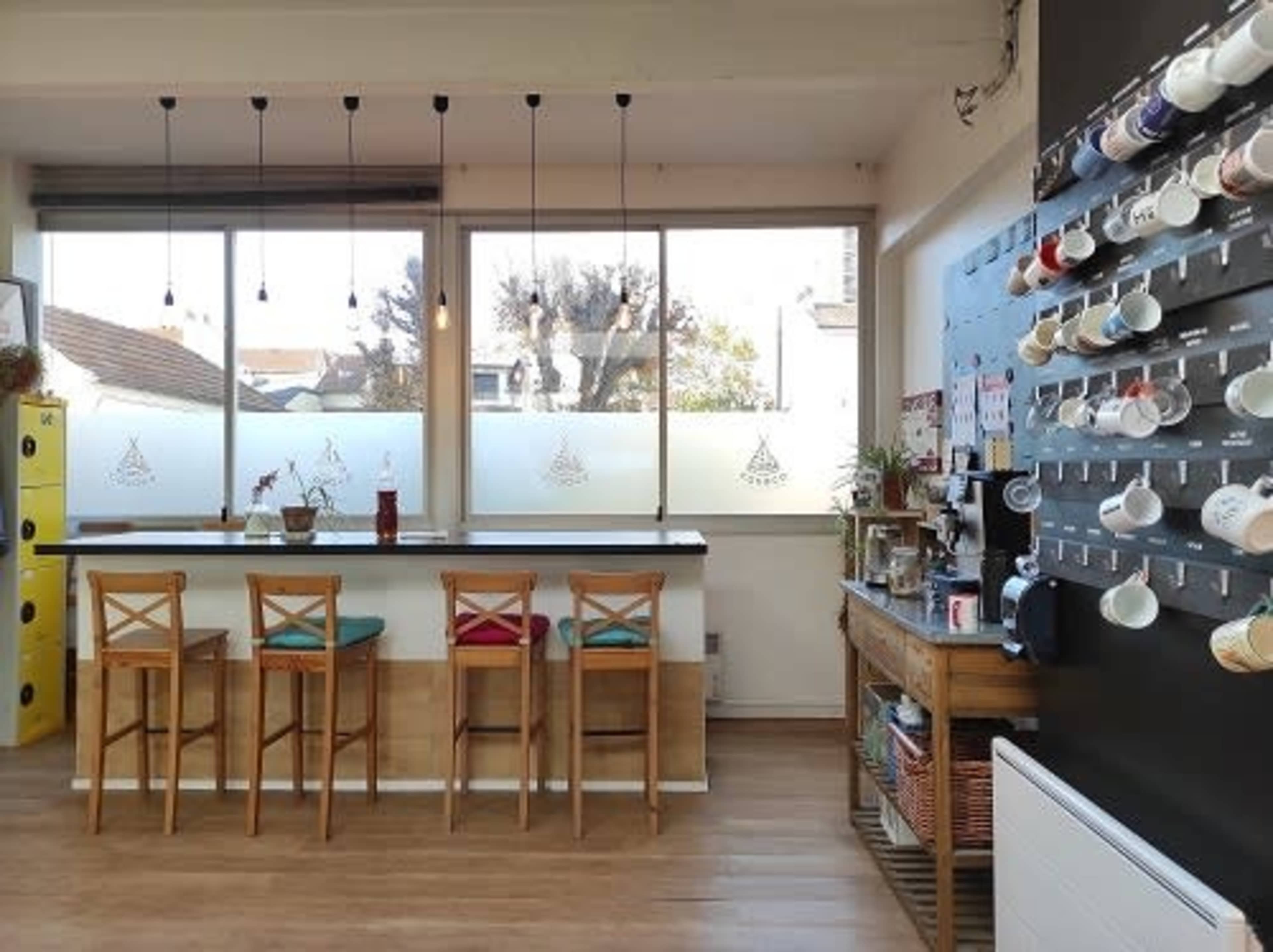 A modern café interior with a counter featuring wooden stools, large windows letting in natural light, and a wall displaying various mugs.