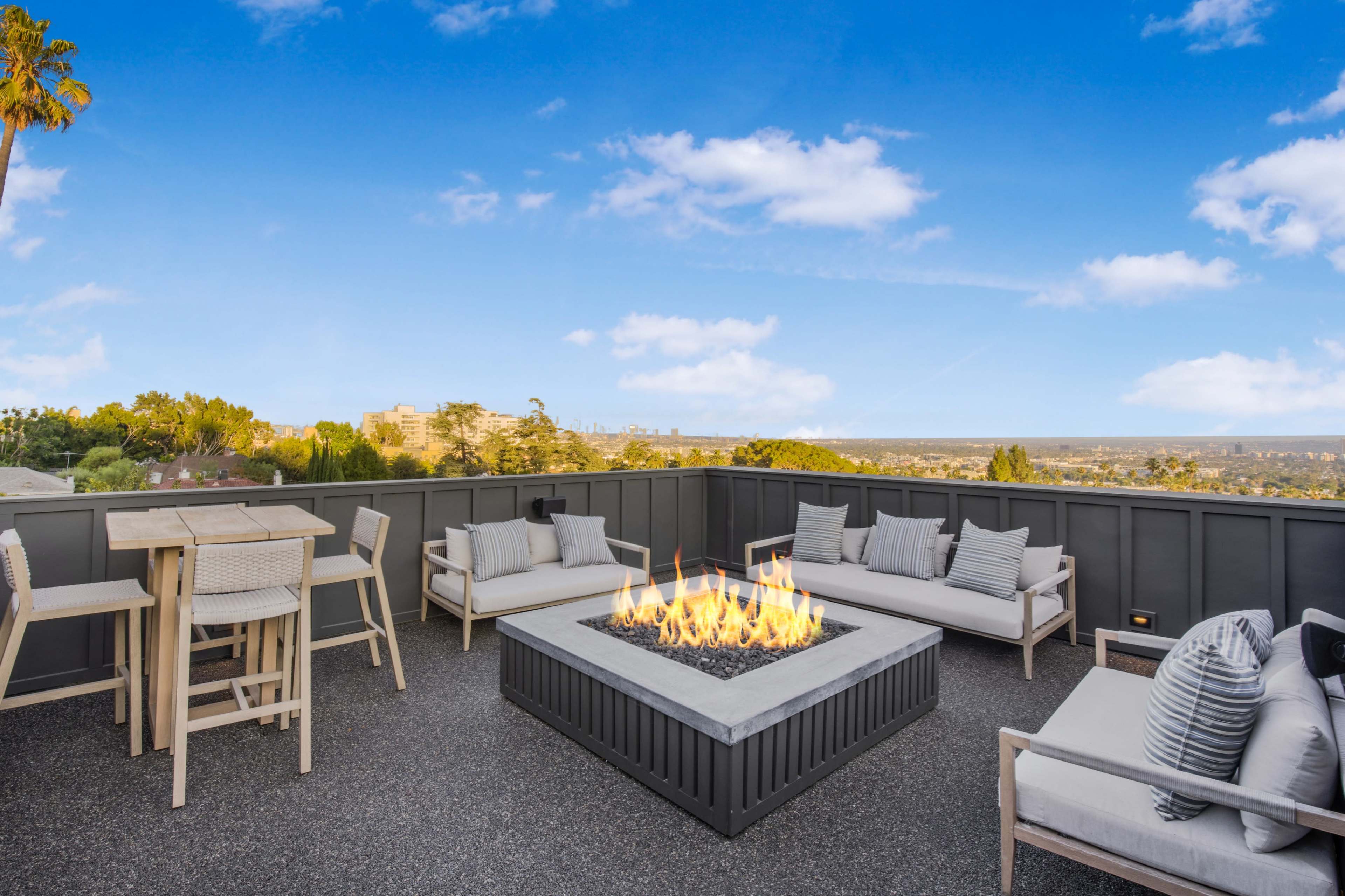The image shows a modern rooftop patio with a fire pit surrounded by seating and a small dining table, overlooking a city skyline and lush greenery.