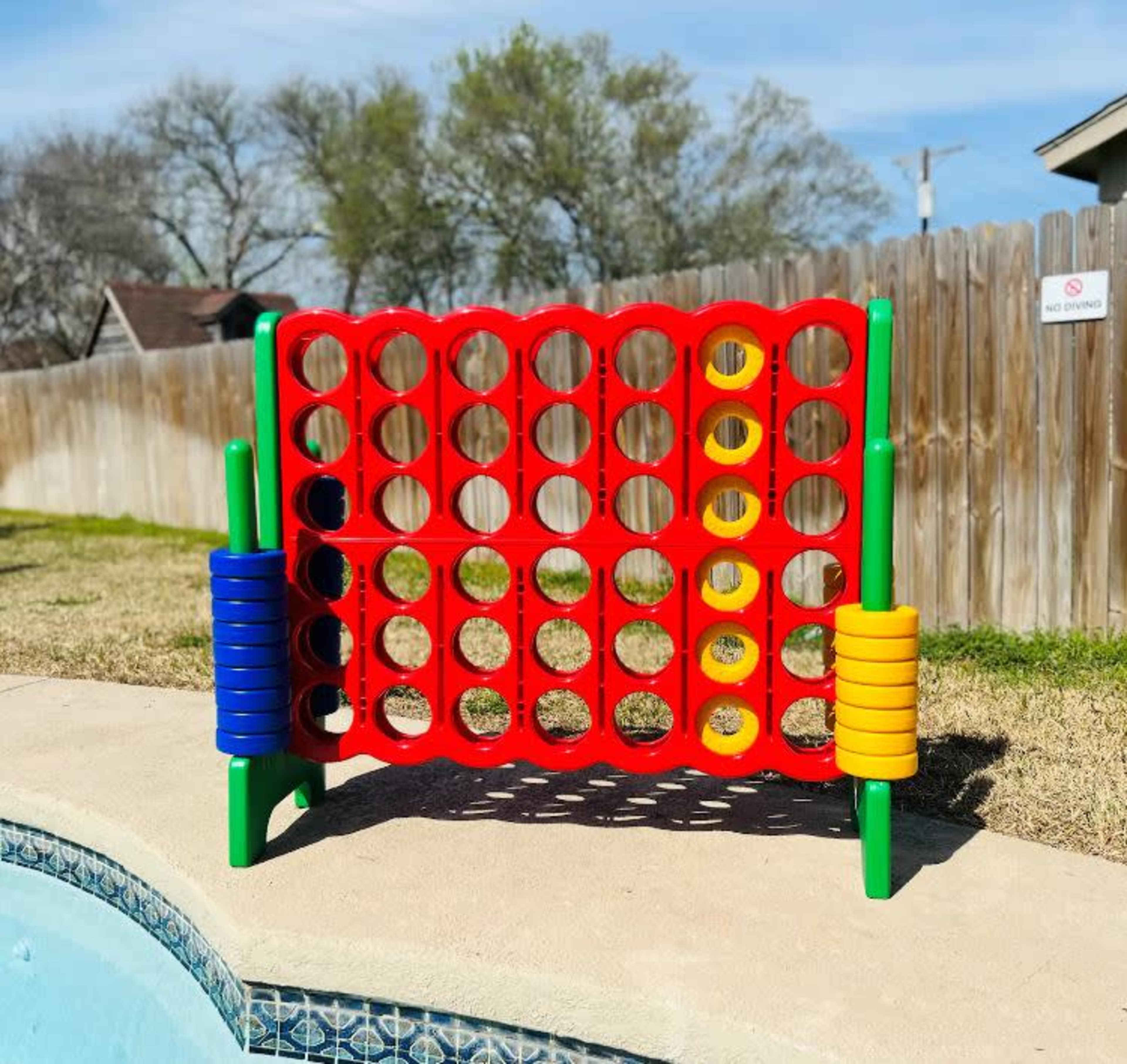 A large red and yellow connect-four game is positioned beside a swimming pool on a grassy area, with a wooden fence in the background.