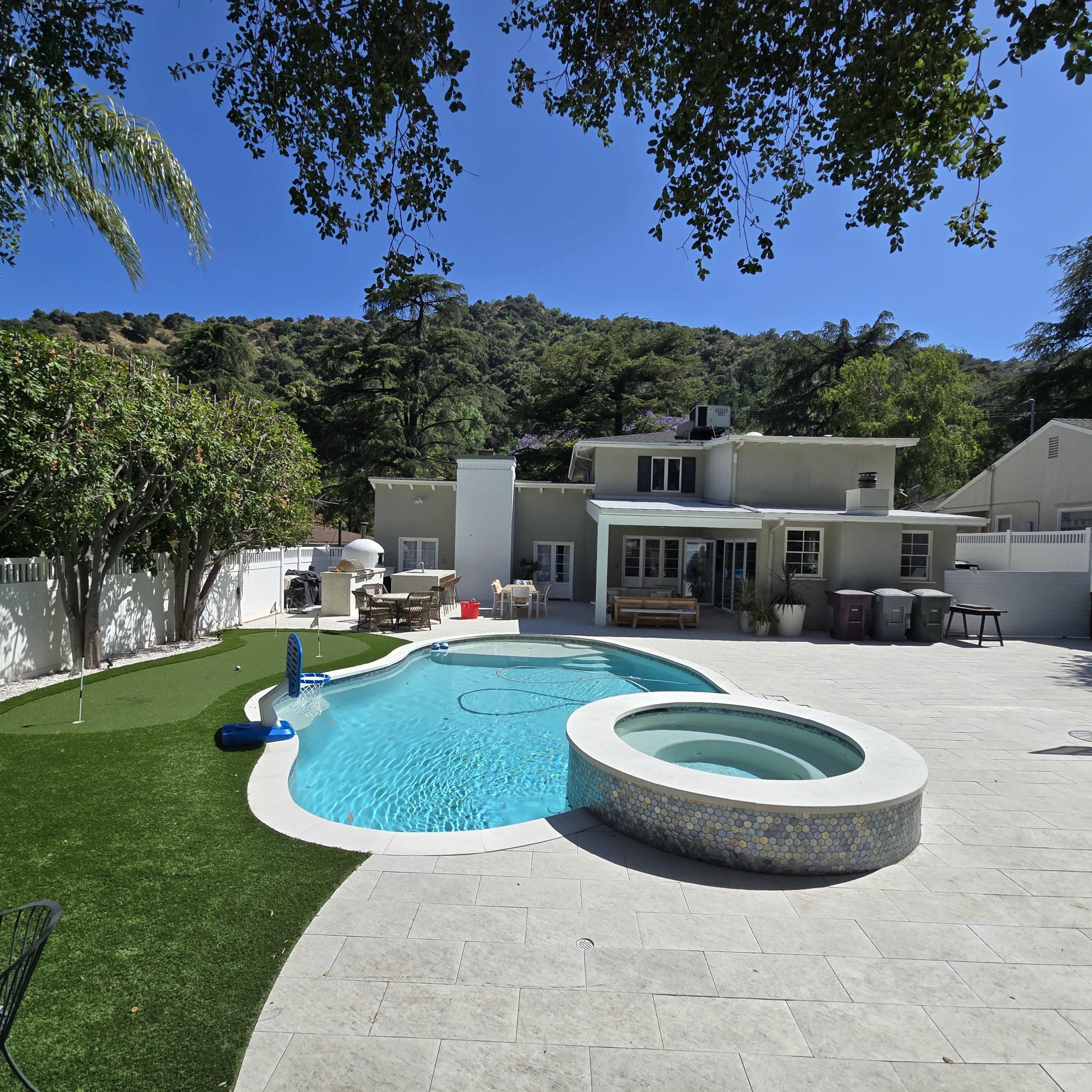 The image shows a residential backyard featuring a swimming pool with a built-in hot tub, surrounded by landscaping and a house in the background.