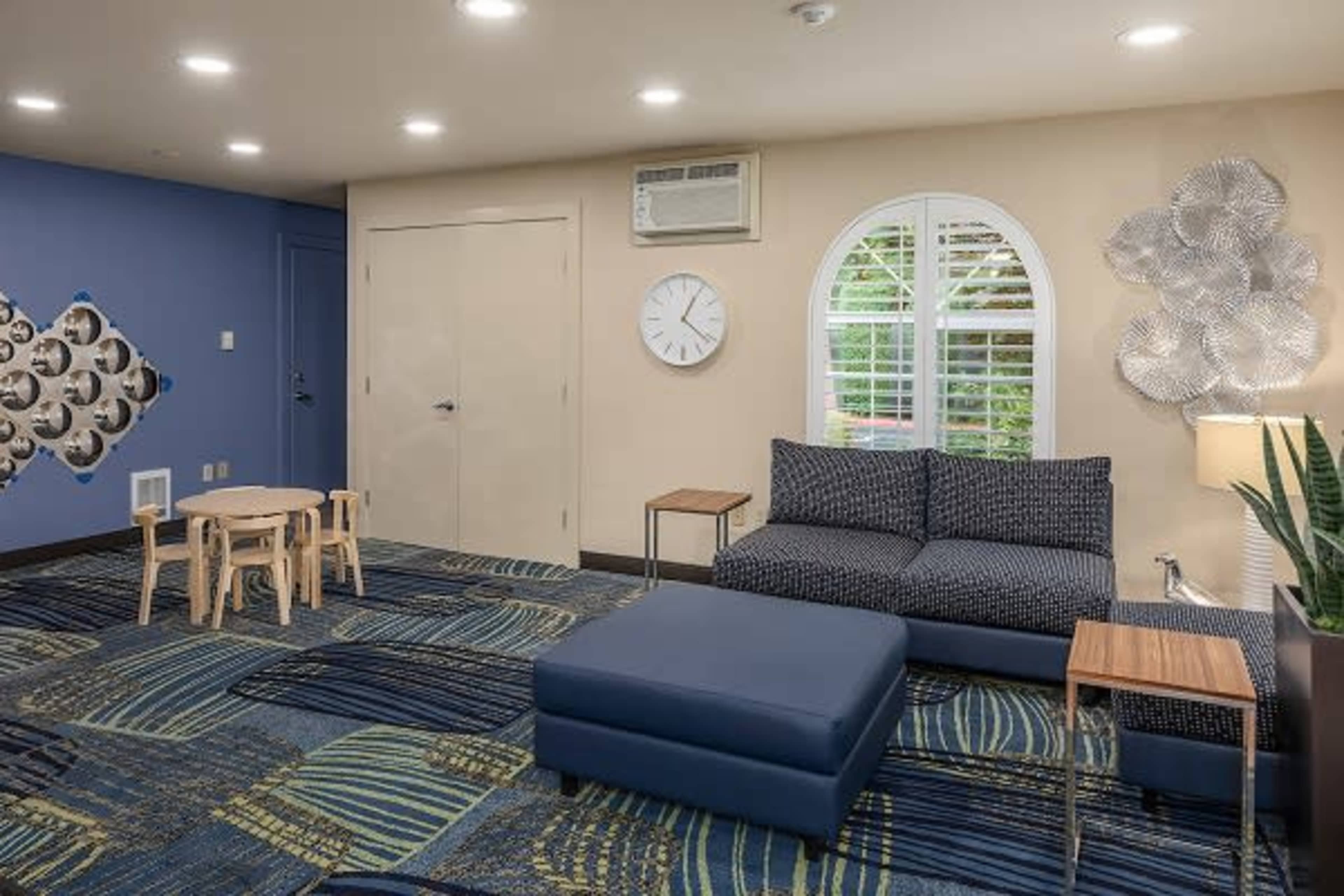 A living space with a blue and beige color scheme, featuring a sofa, a small table with chairs, a wall clock, and decorative elements.