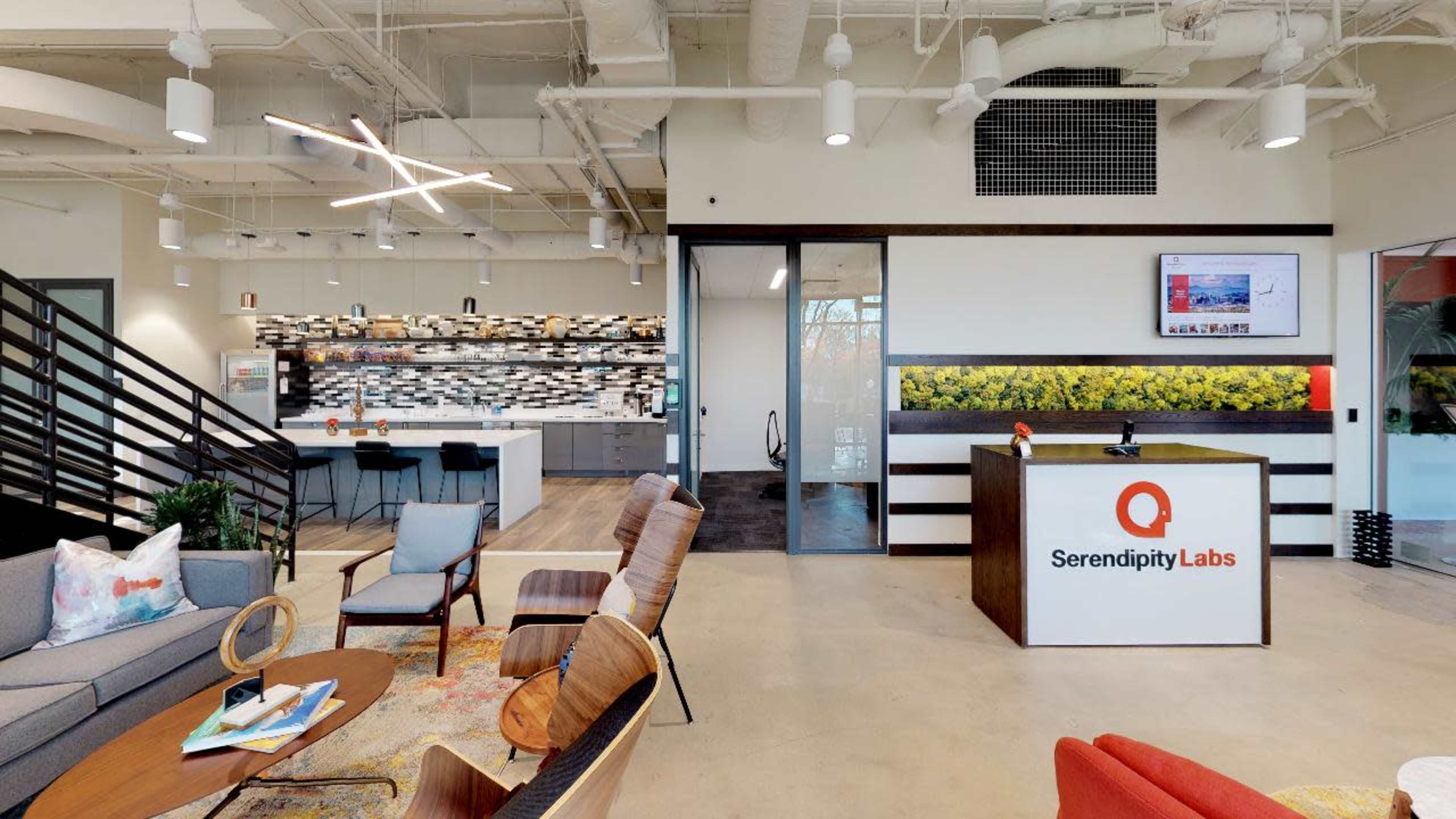 The image shows a modern office reception area with a front desk labeled "Serendipity Labs," surrounded by seating and an open kitchen space.