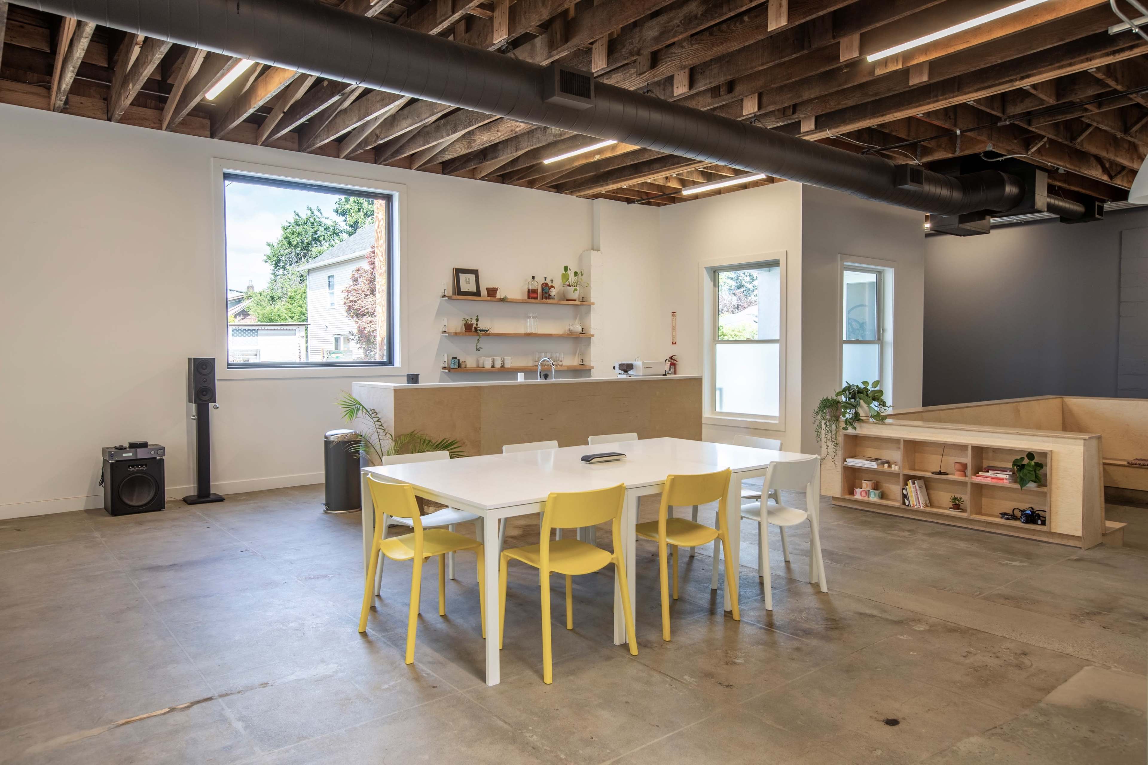 A spacious, modern interior with a white dining table surrounded by yellow and white chairs, hardwood beams overhead, and a corner featuring a low bookshelf and seating area.