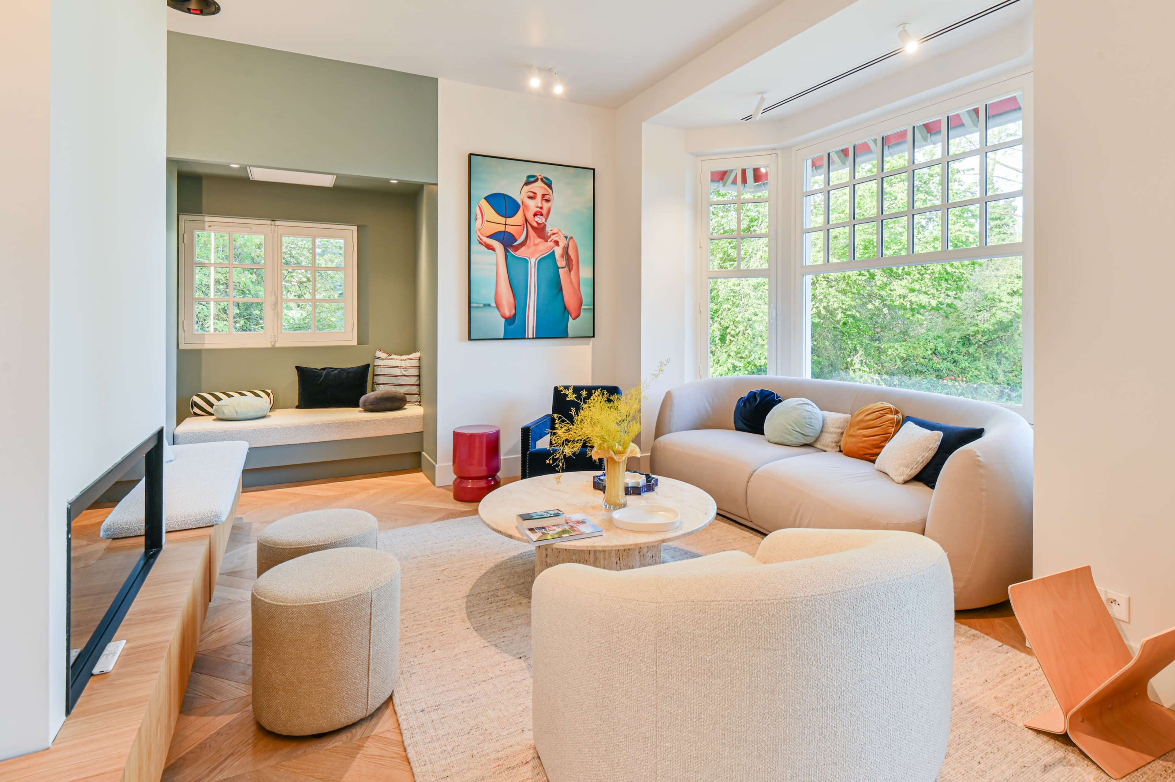 The image depicts a modern living room featuring a curved sofa, circular coffee table, ottomans, a large artwork on the wall, and a cozy reading nook with windows.