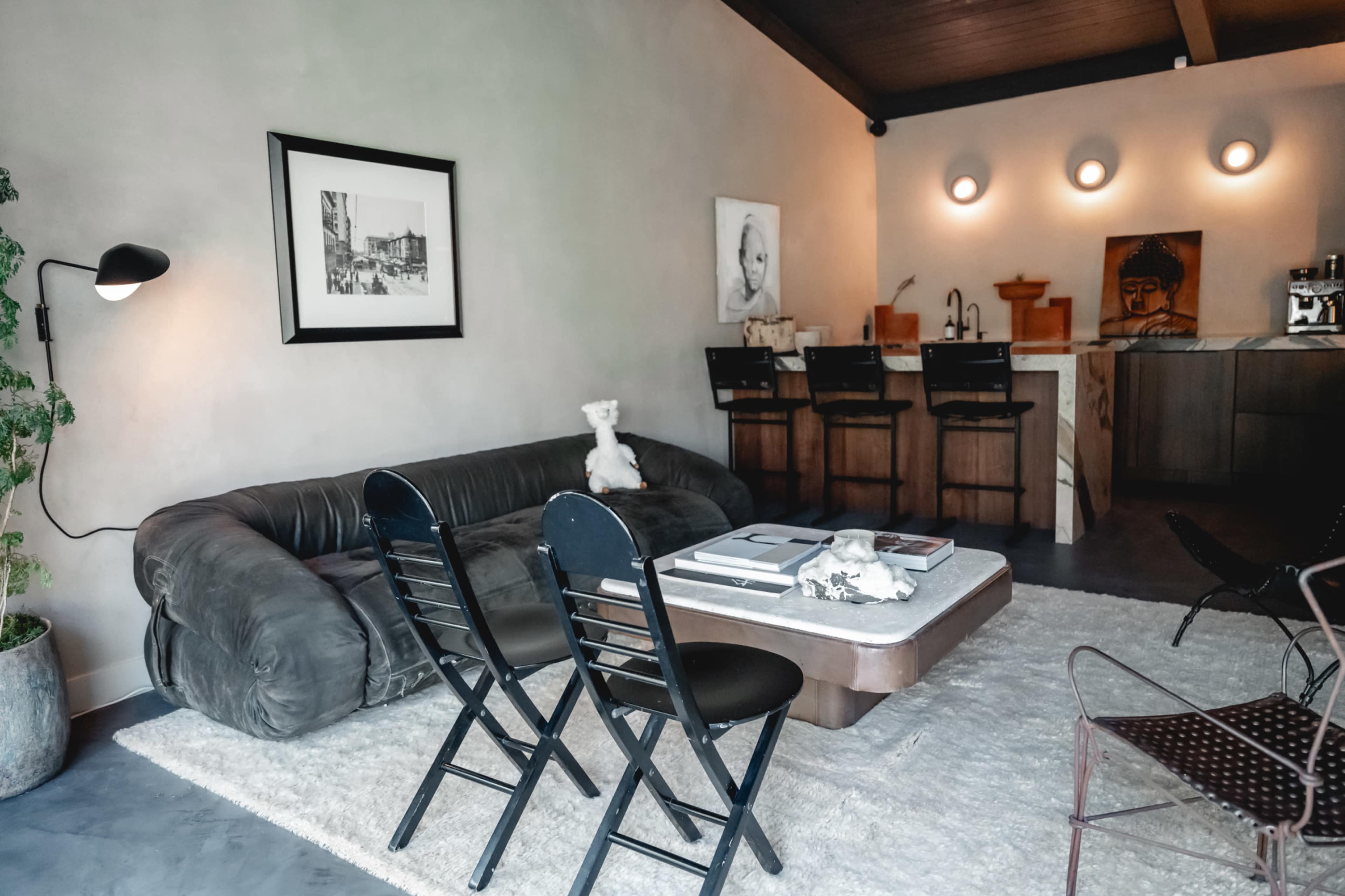 A modern living room featuring a dark leather sofa, a coffee table, and a small dining area with four barstools against a wall, all set on a light rug.